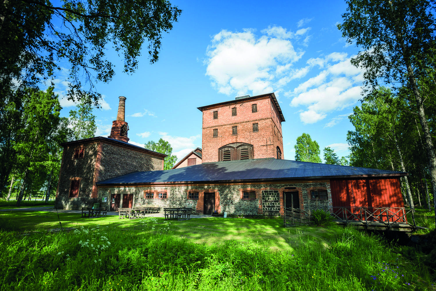 Ein altes Gebäude mit Schornsteinen aus Ziegeln ist von viel Grün umgeben.