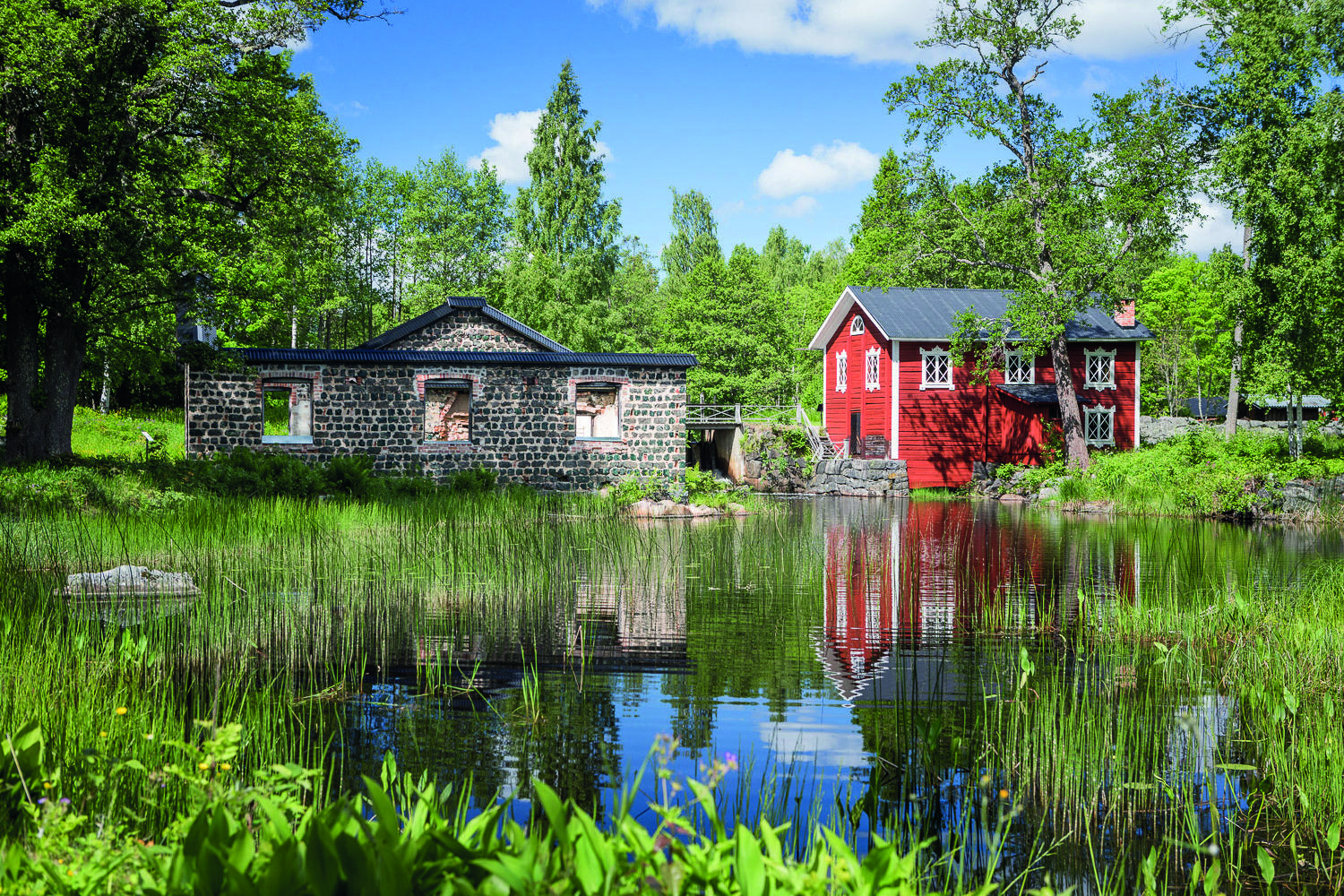 Ein rotes Holzhaus mit weißen Zierleisten und ein altes Steinhaus neben einem See im Sommer.