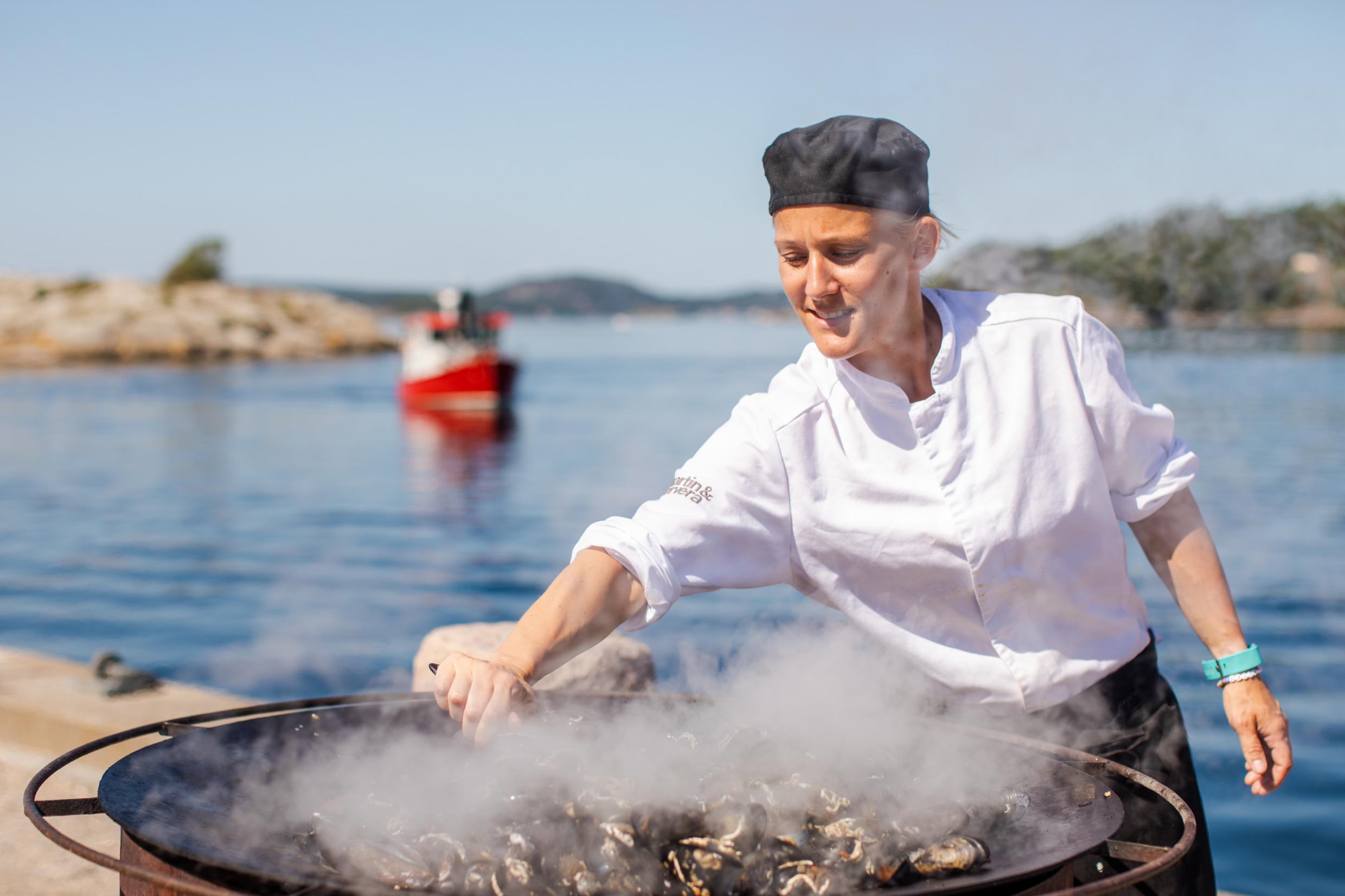 Ein Koch steht draußen am Meer und kocht Muscheln in einer großen Pfanne.