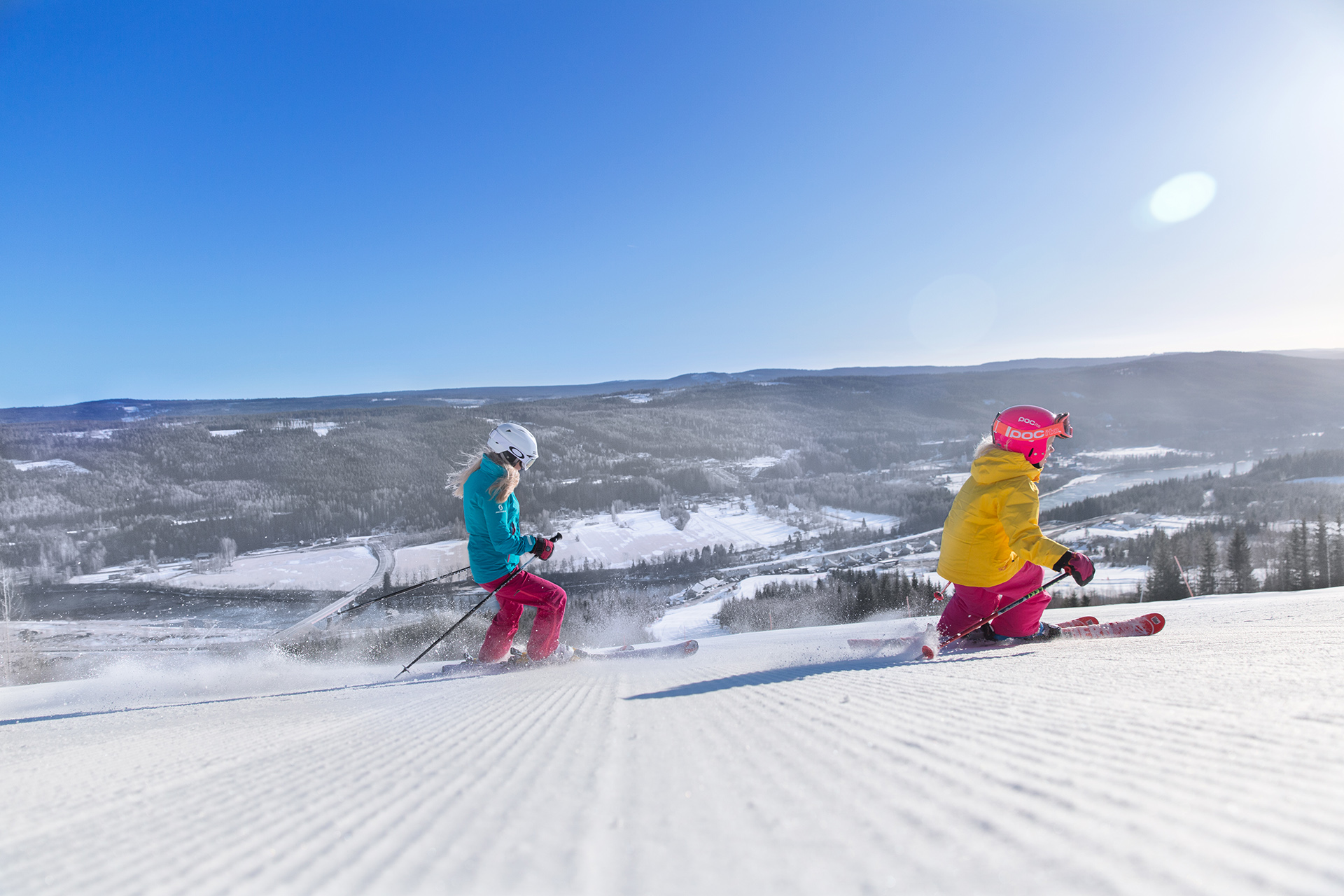 Two skiers on a freshly groomed slope in Branäs, with forested hills and snowy landscape in the background.