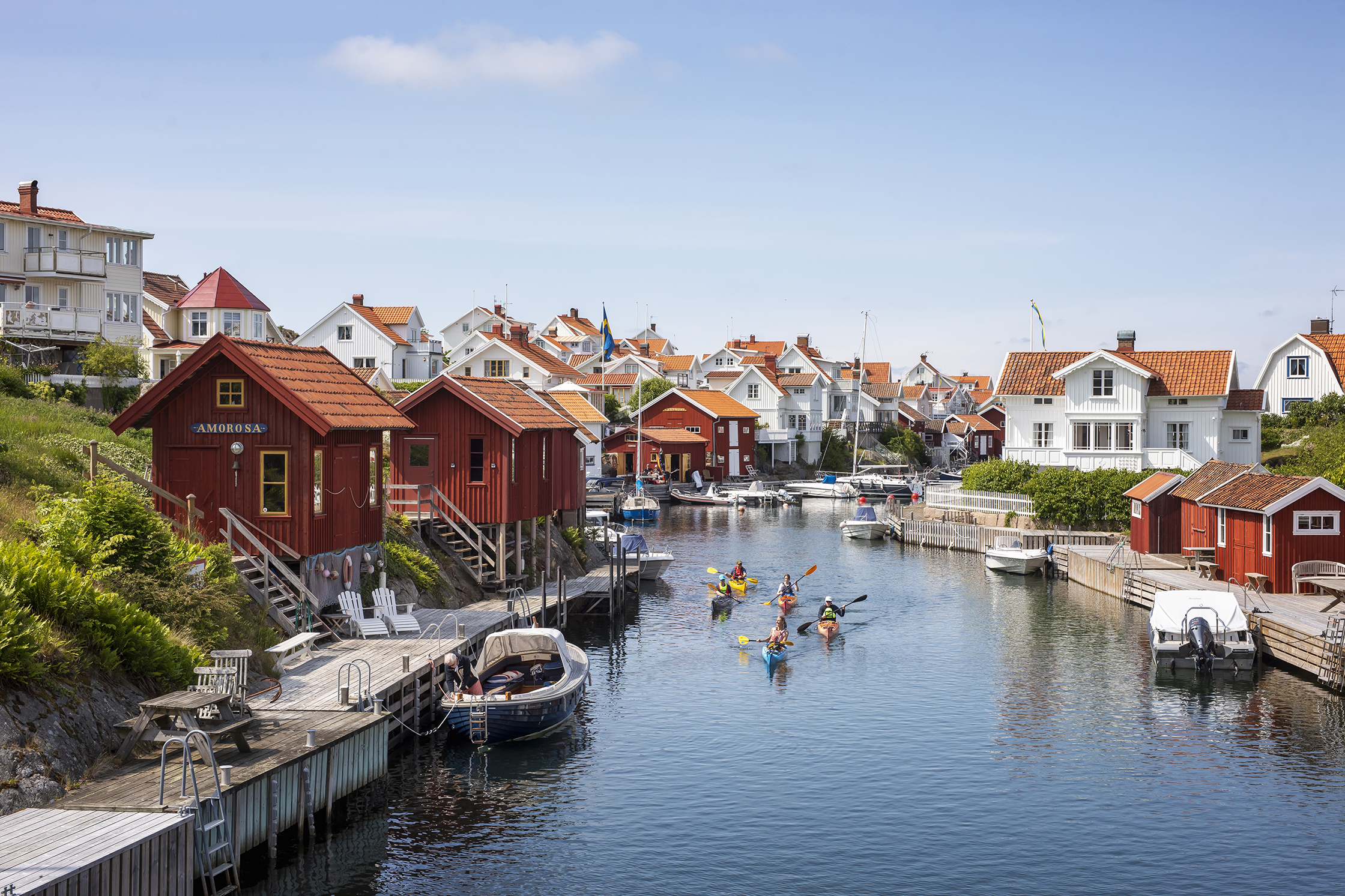 Kajakkers peddelen door een smalle waterweg in Grundsund, omringd door rode boothuizen, witte houten huizen en aangemeerde boten.