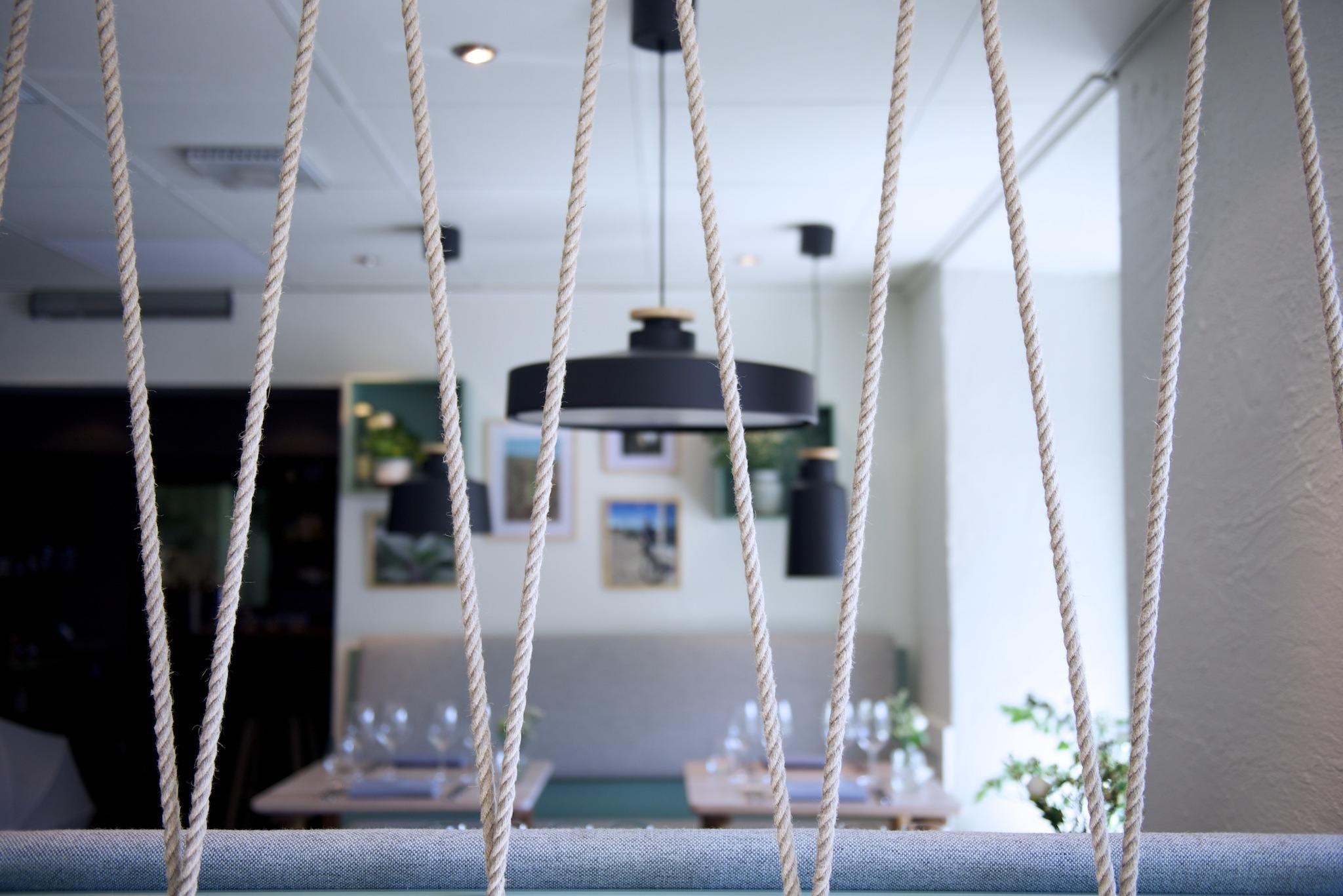 Innenraum des Restaurants Bhoga in Göteborg. Hinter Seilen, die als Dekoration aufgestellt sind, sieht man im Hintergrund zwei gedeckte Tische verschwommen.