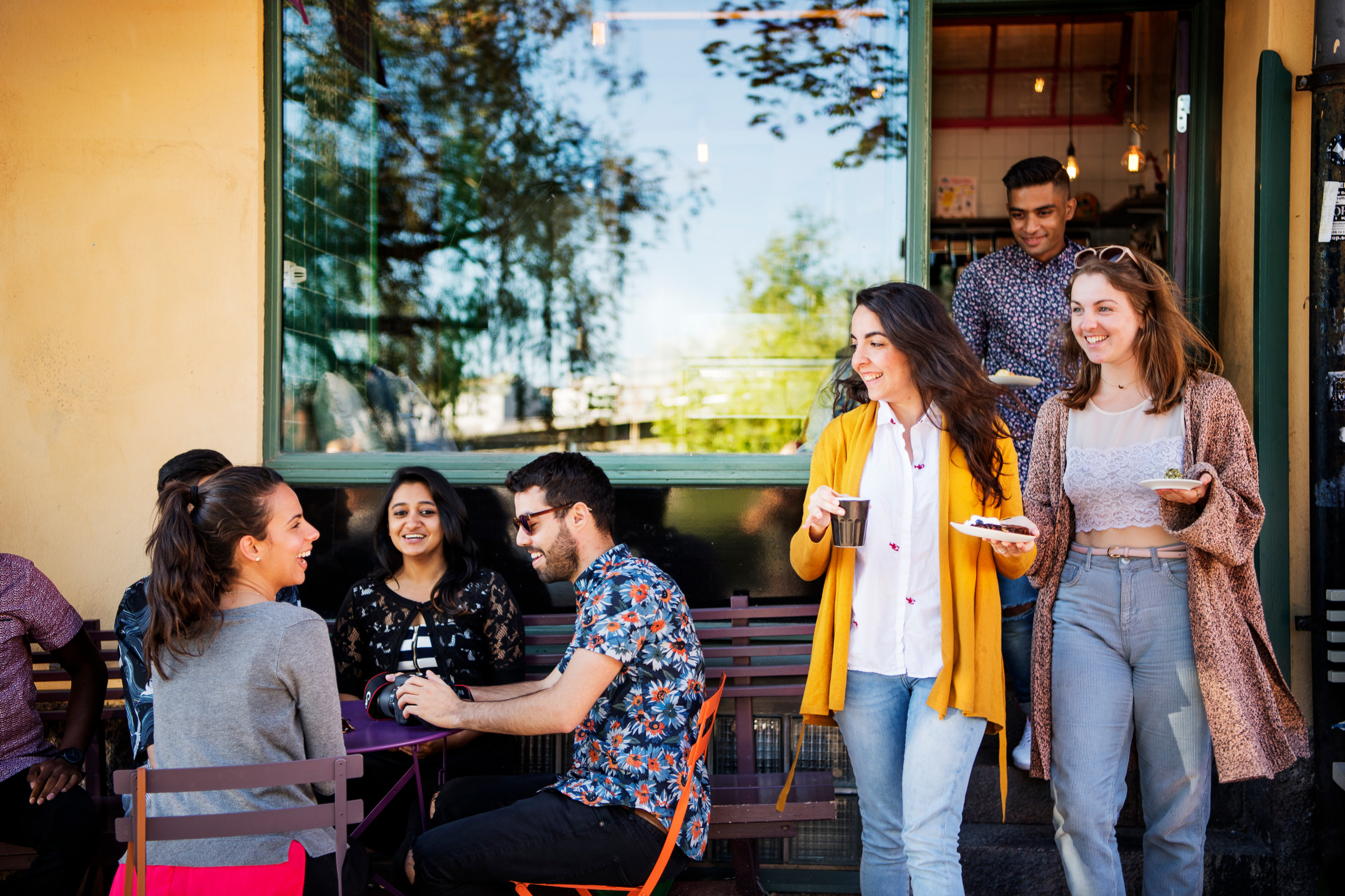 Mensen komen uit een café met koffie en snoepjes. Het terras zit vol.