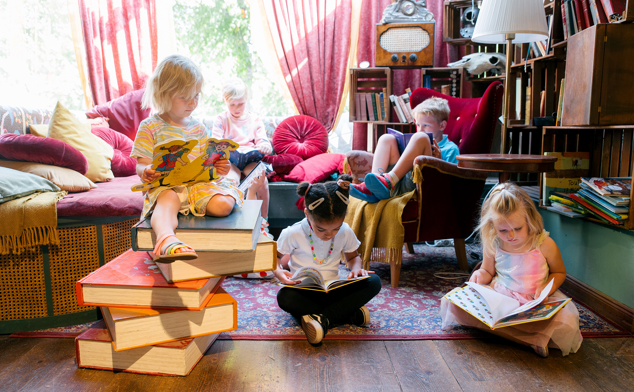 Children reading books in a colourful, cosy room at Junibacken, surrounded by oversized books, cushions and armchairs.