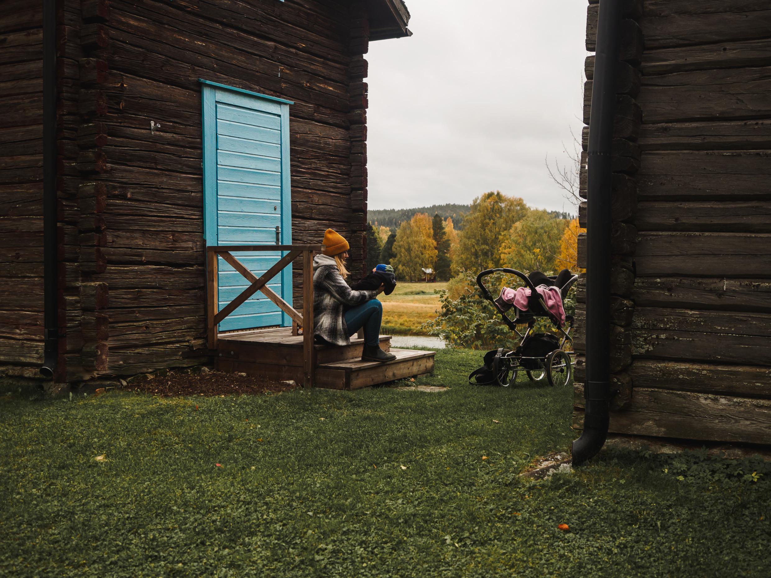 Eine Frau sitzt mit einem Baby auf einer hölzernen Veranda. Der Kinderwagen steht daneben.