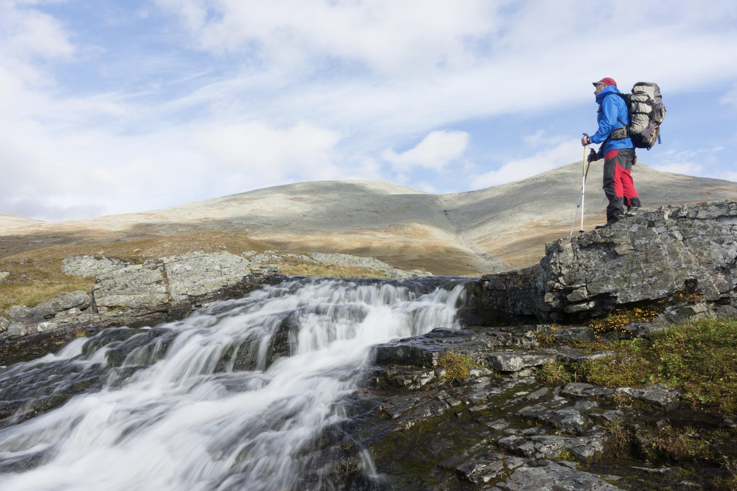 Ein Mann mit Wanderausrüstung steht neben einem kleinen Fluss mit Stromschnellen.