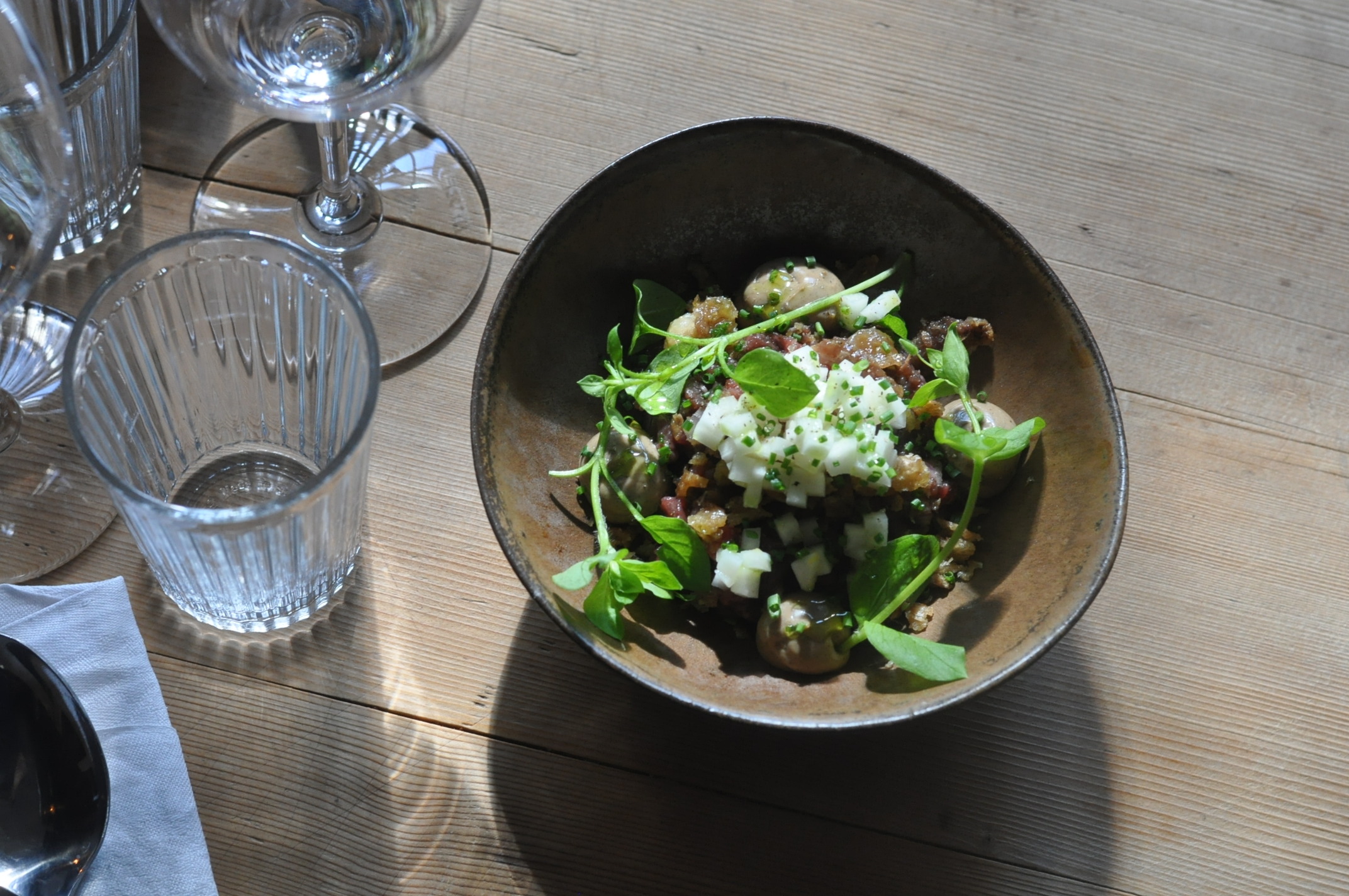 Ein nordisches Gericht mit fein geschnittenem Gemüse, Blattgemüse und Pilzen im Restaurant Bua in Östersund, serviert auf einem Holztisch mit Gläsern.
