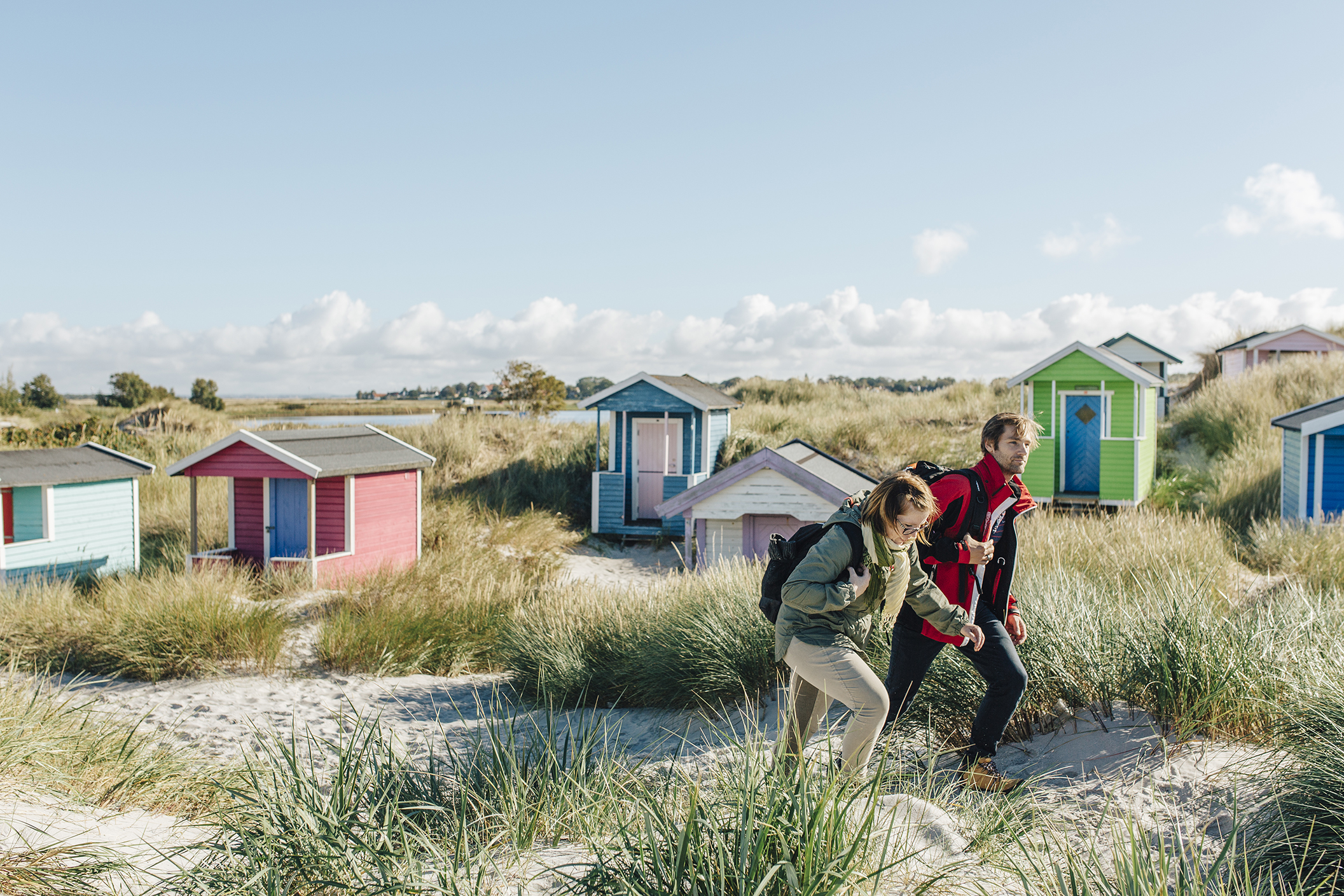 Zwei Wanderer spazieren zwischen Sanddünen und bunten Strandhütten in Küstennähe in Skåne.