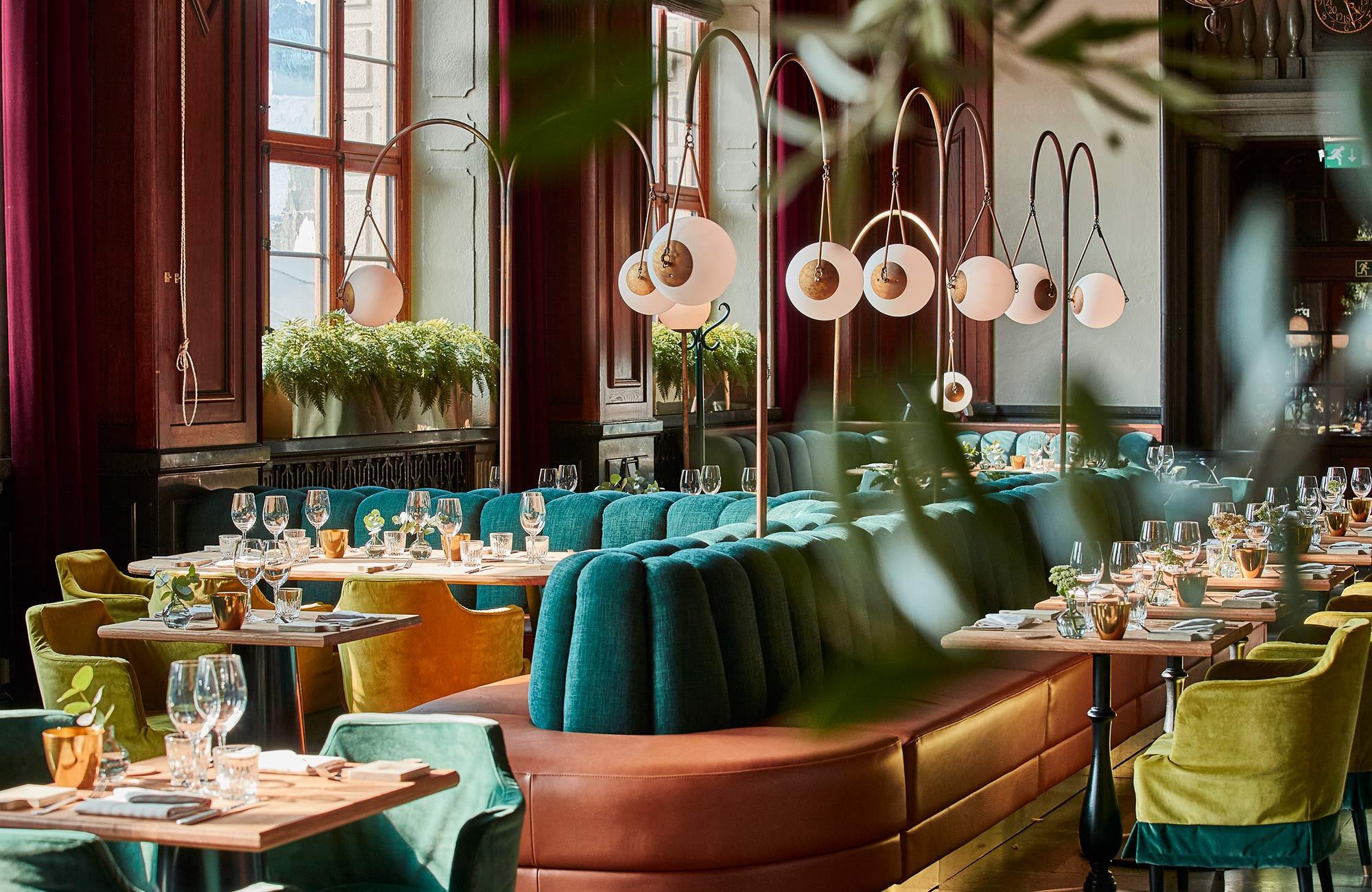 Een restaurant met fluwelen meubels en bruin leer. De tafels zijn gedekt met glazen, bestek en servetten.