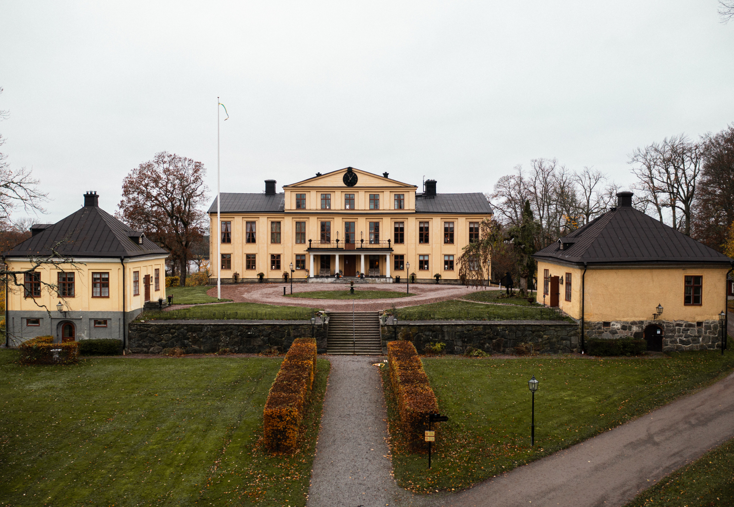 Vooraanzicht van Krusenberg Manor in Knivsta, een groot geel landhuis uit de 17e eeuw, omringd door bomen en tuinpaden op een bewolkte herfstdag.