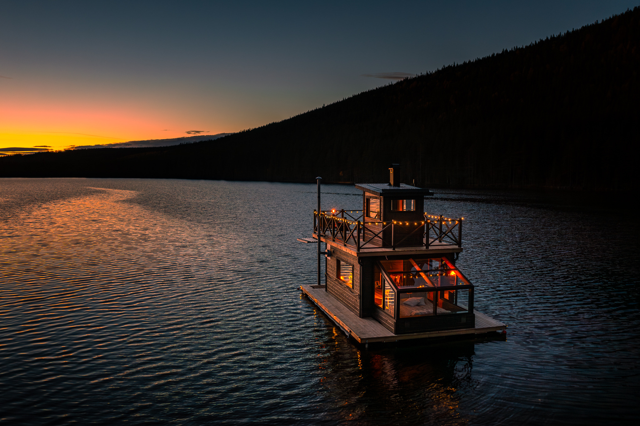 La cabane flottante Raven sur un lac calme au coucher du soleil, avec des lumières scintillantes et des collines boisées en arrière-plan.