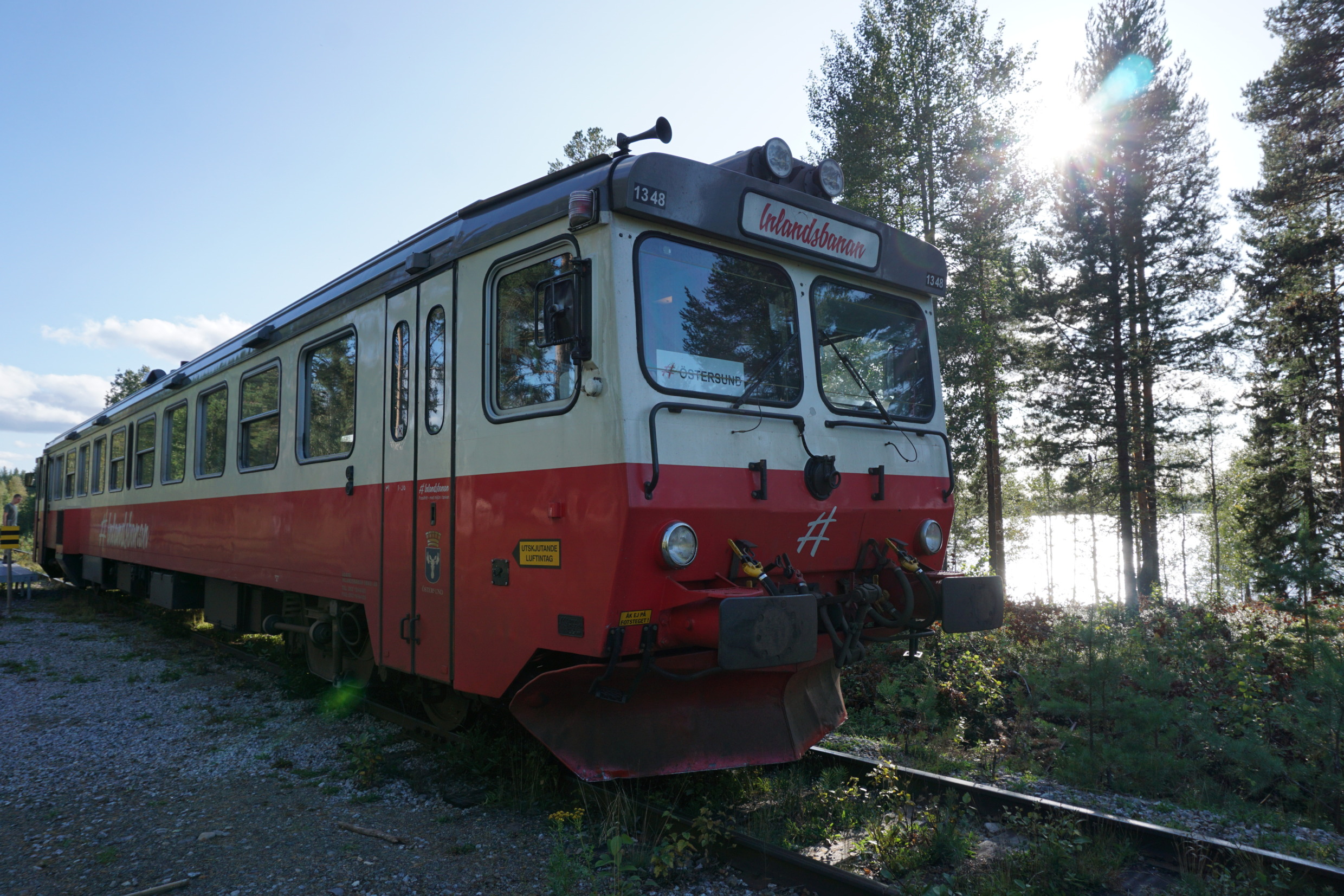 A red and white train standing still on a railway. Behind it there is a lake and tall trees.