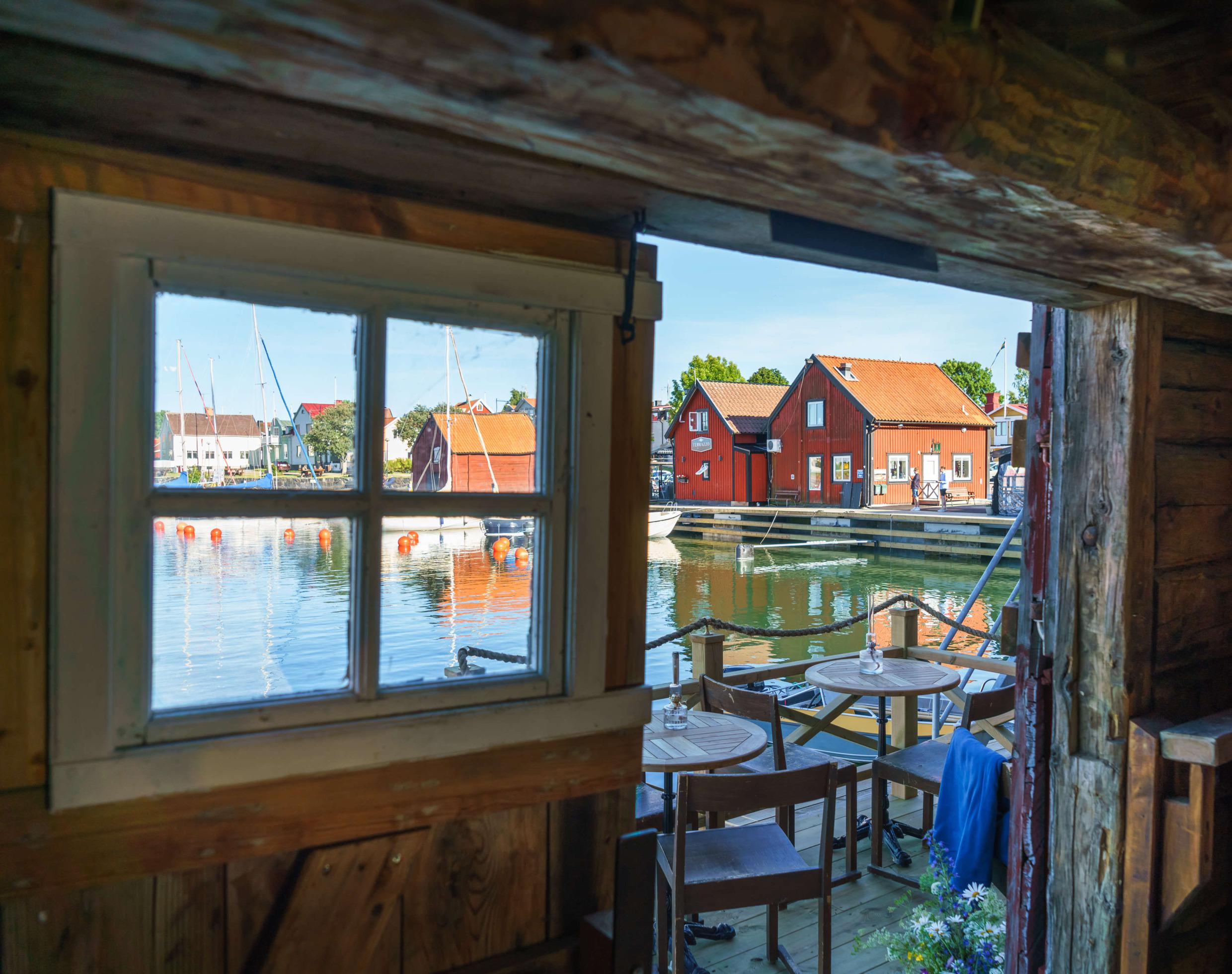 Vue à travers une porte depuis l'intérieur d'un restaurant. Il y a une petite terrasse extérieure en bois avec quelques tables et chaises au bord de la mer. Une marina et plusieurs chalets se trouvent en arrière-plan.