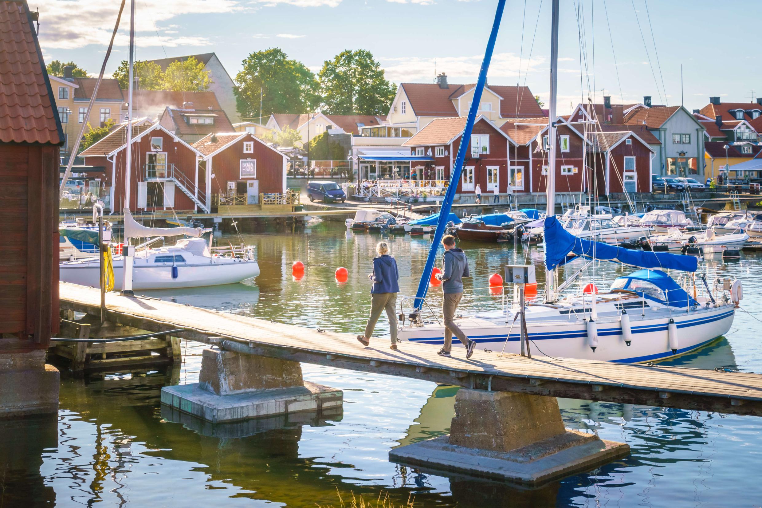 Een stel loopt op een steiger in een jachthaven in een kleine houten stad. Langs het water staan rode huisjes.