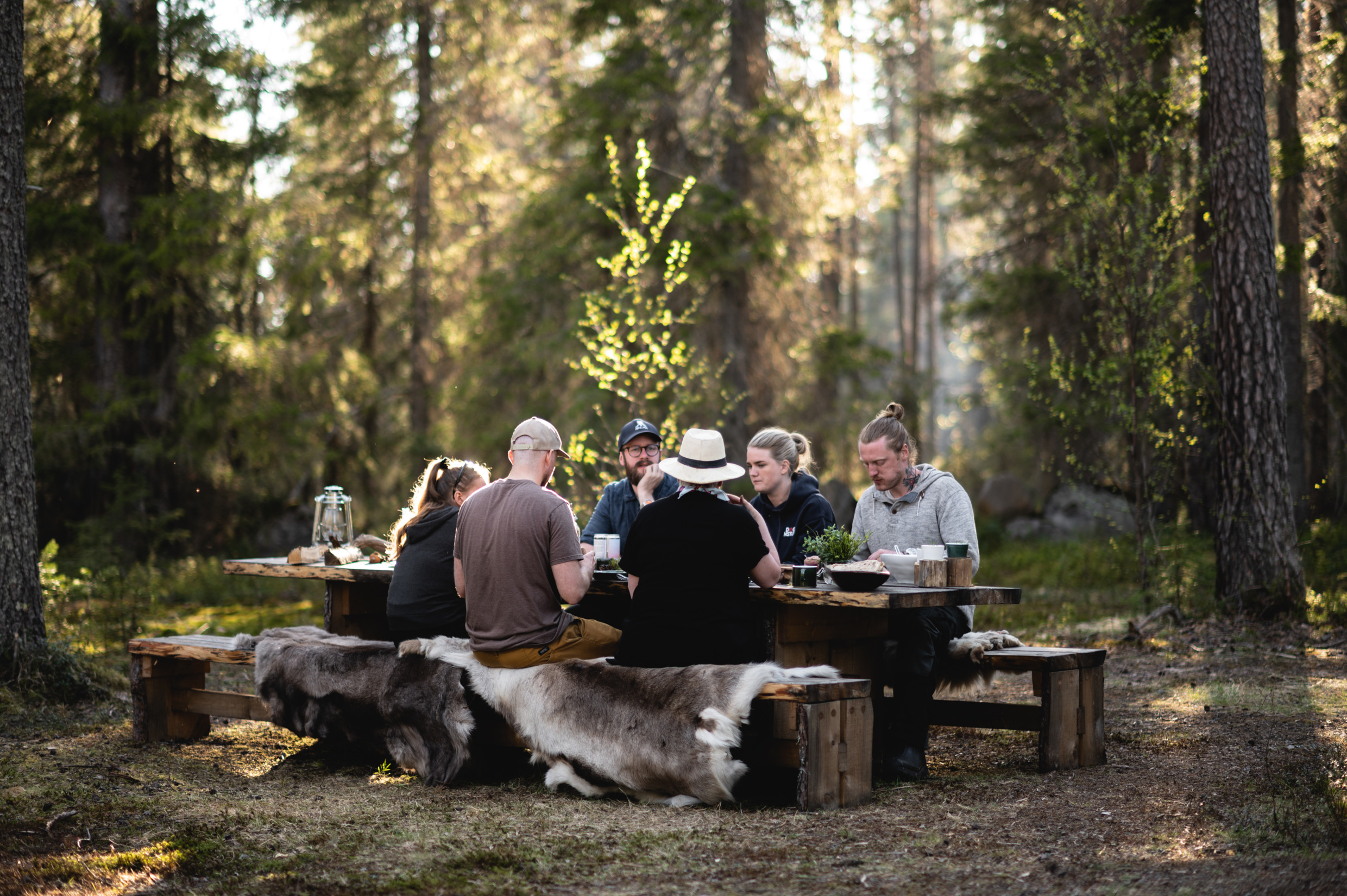 Six personnes assises à une table en bois dans une forêt. Elles sont assises sur des bancs recouverts de peaux de mouton. Le soleil brille à travers les arbres.