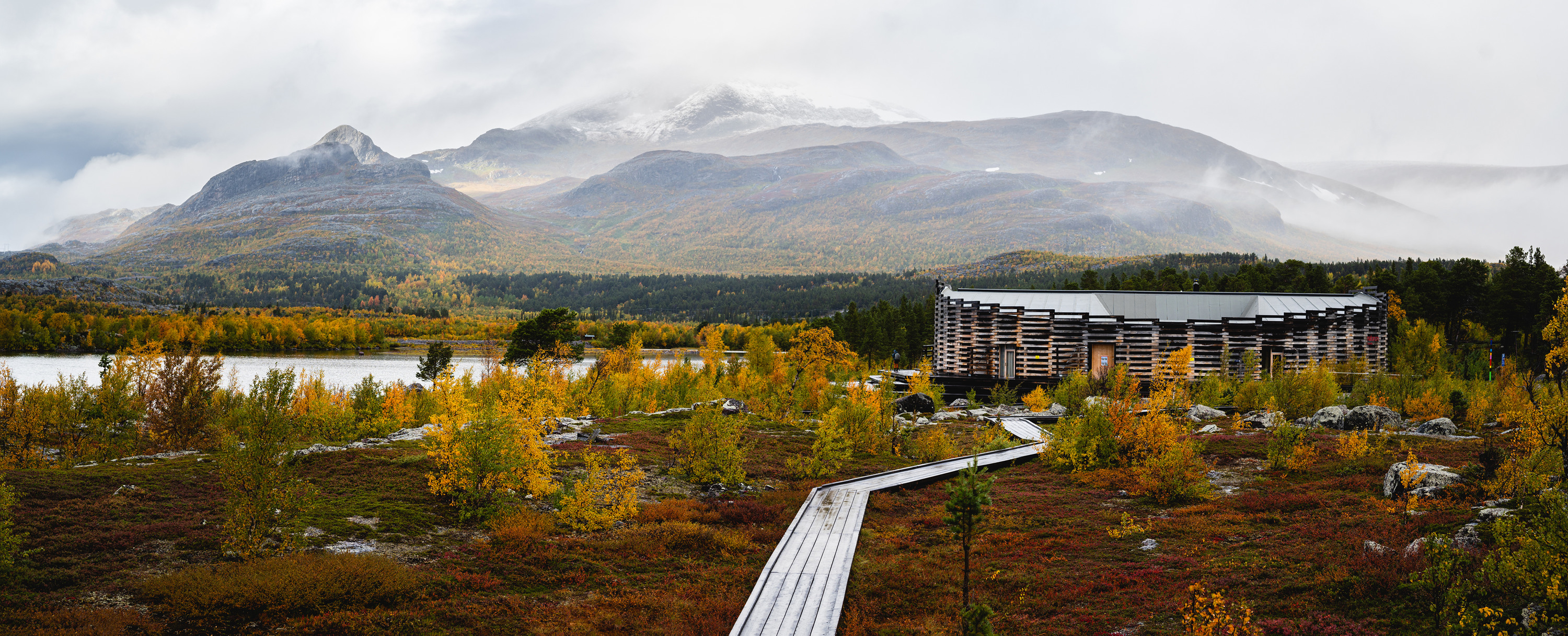 A modern wooden visitor centre stands beside a boardwalk overlooking colourful autumn vegetation, a lake and snow-dusted mountains.