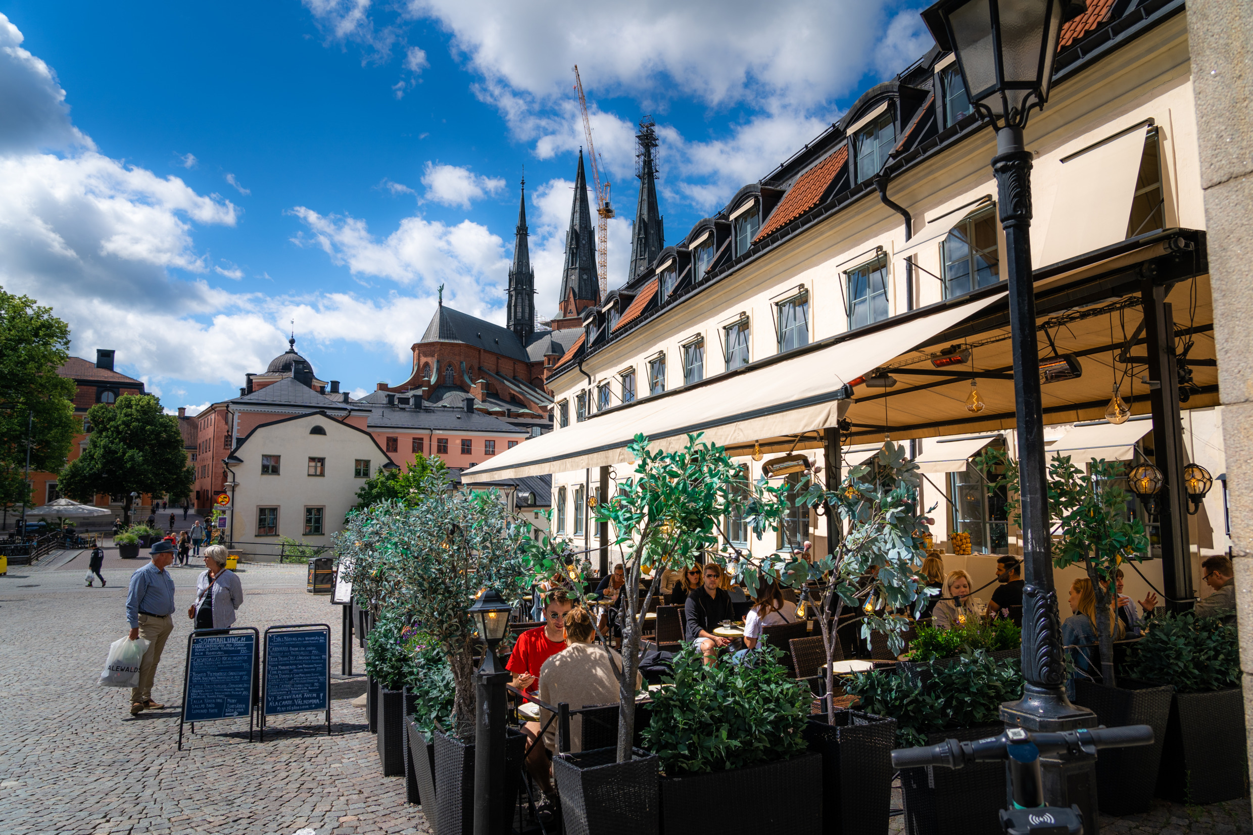 Zitgedeelte buiten met mensen die eten en drinken in de zon, vlakbij de kathedraal van Uppsala. Beige huis en een plein met geplaveide straatjes.