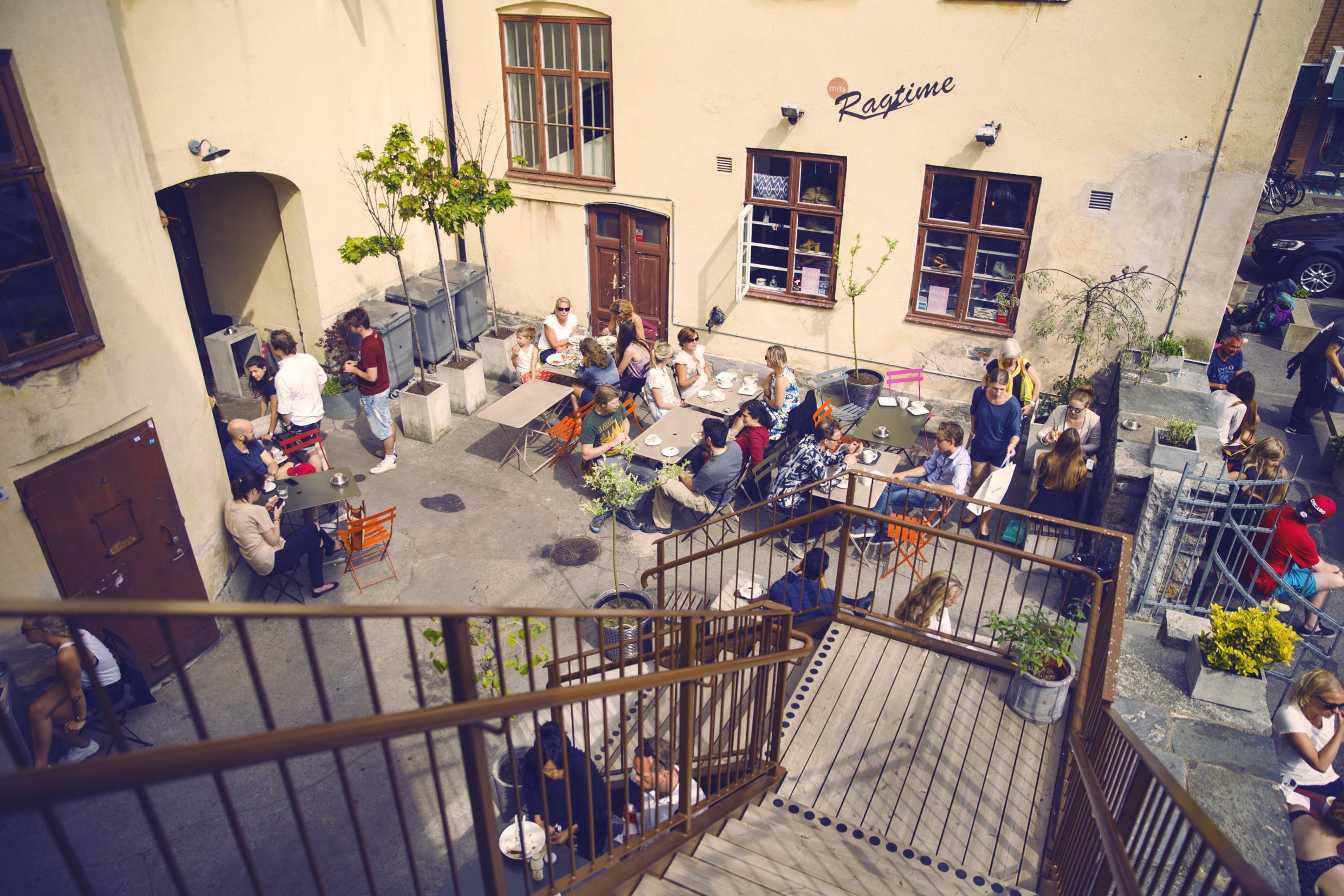Des personnes sont assises à des tables dans la cour d'un café. En arrière-plan, il y a une maison de couleur jaune pâle sur laquelle est écrit "Ragtime".