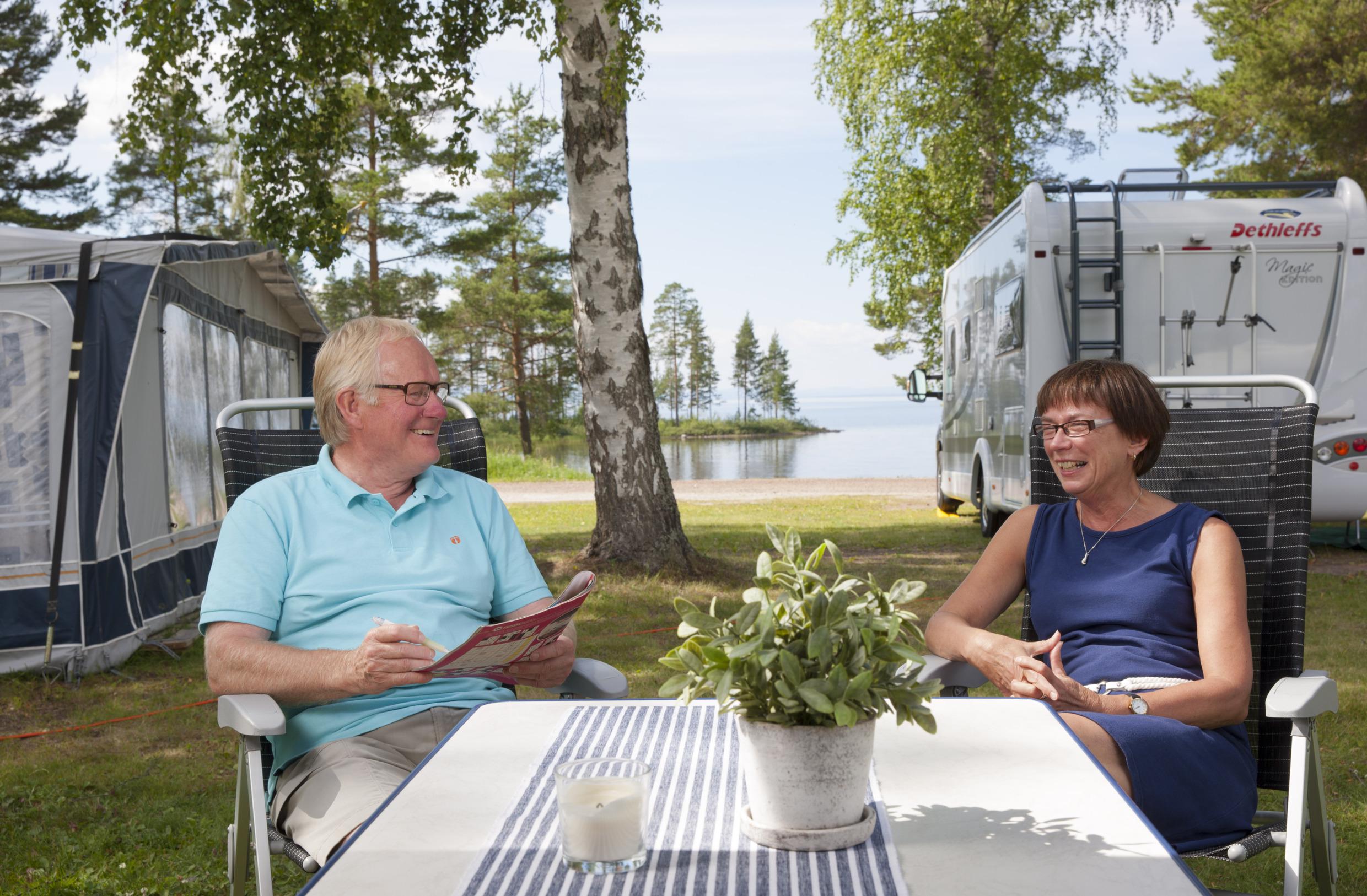 Op een camping zit een stel aan een tafel. Je ziet achter hen een tent en een caravan en op de achtergrond een meer.