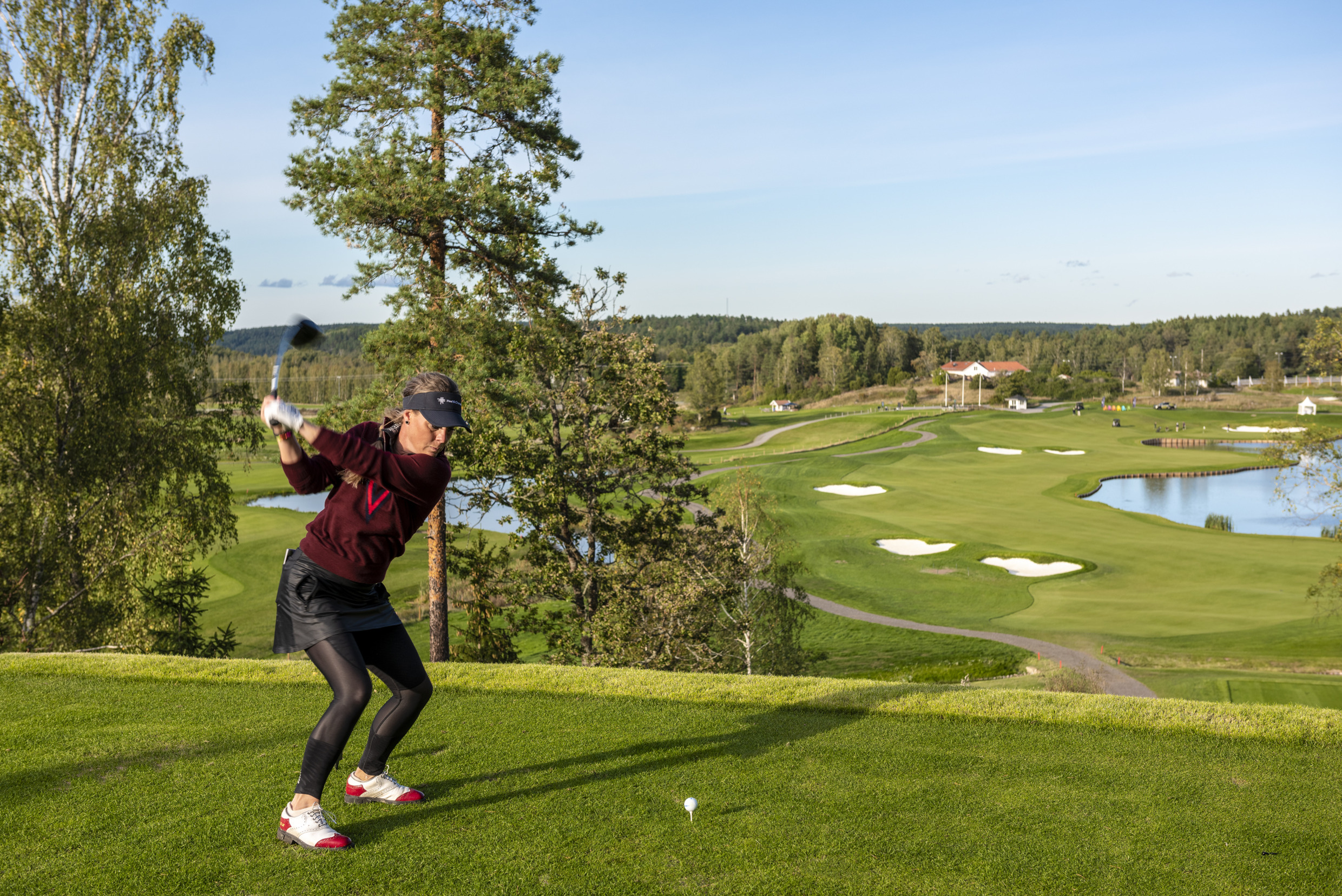 Female golfer dressed in black and red clothing and a golf cap playing golf. In the background, the golf course, a small lake, and a house.