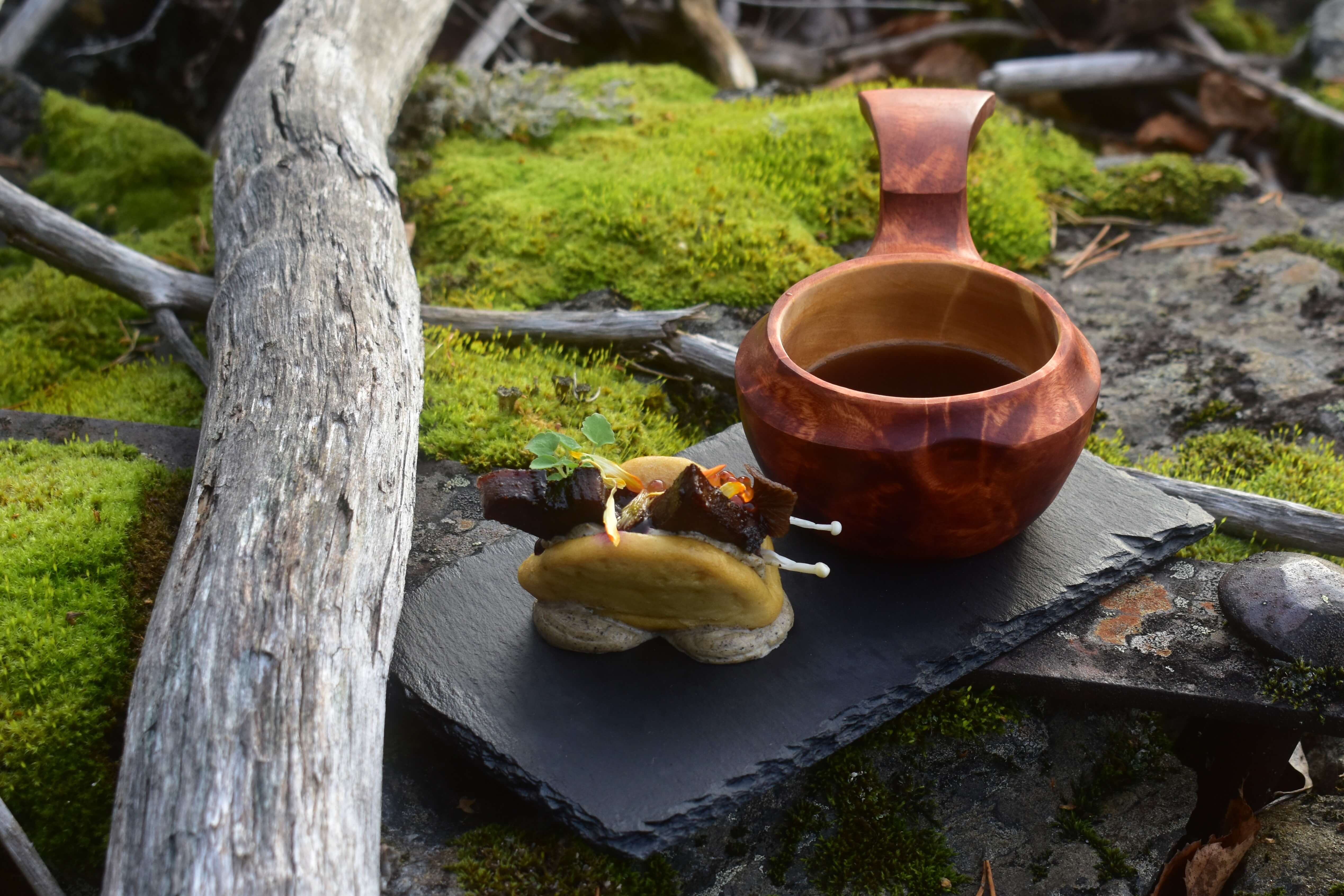 A gourmet dish and a wooden cup served on a slate plate among moss-covered rocks at Wild River near Umeå.