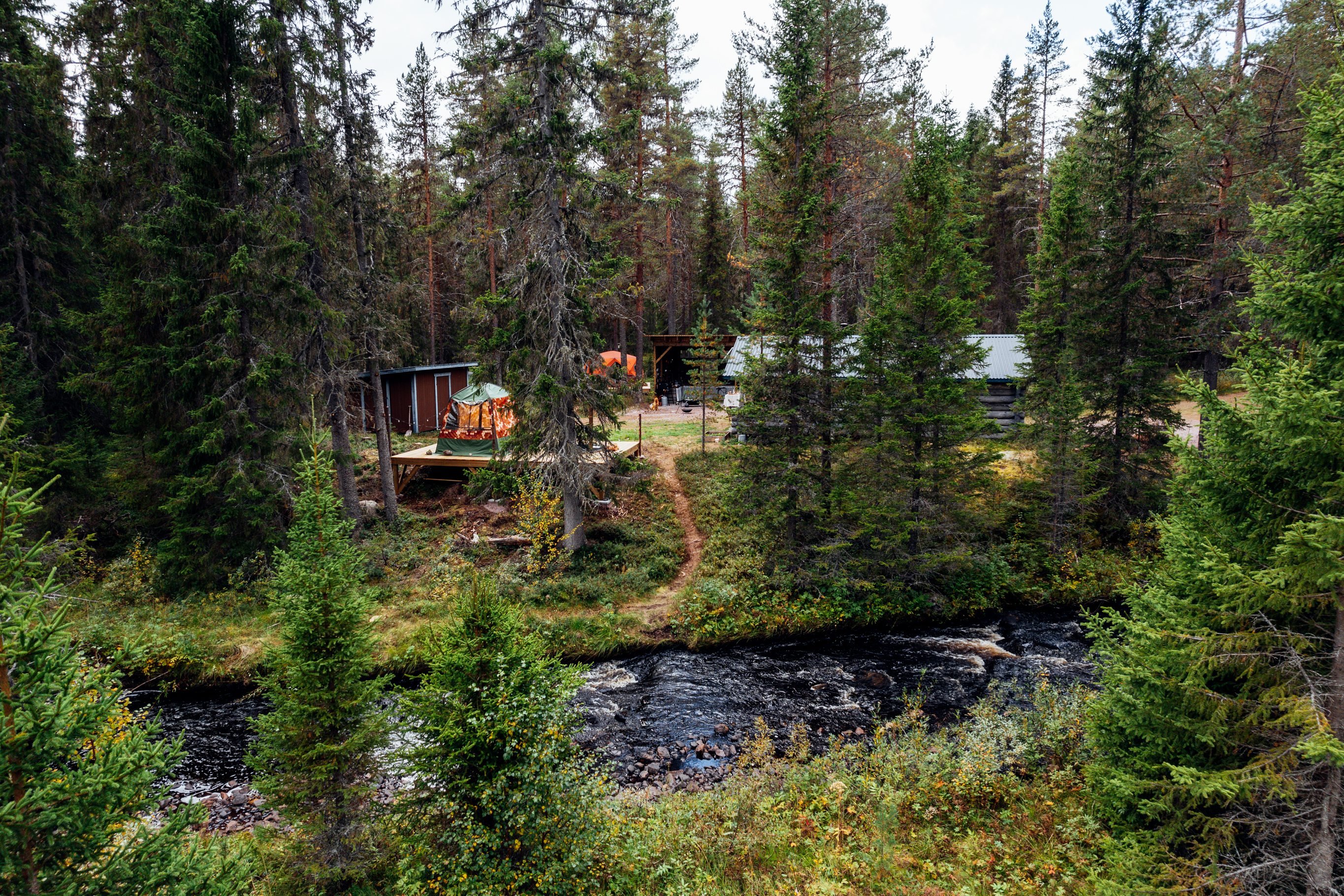 A forest camp with tents and wooden structures set beside a river, surrounded by dense trees.