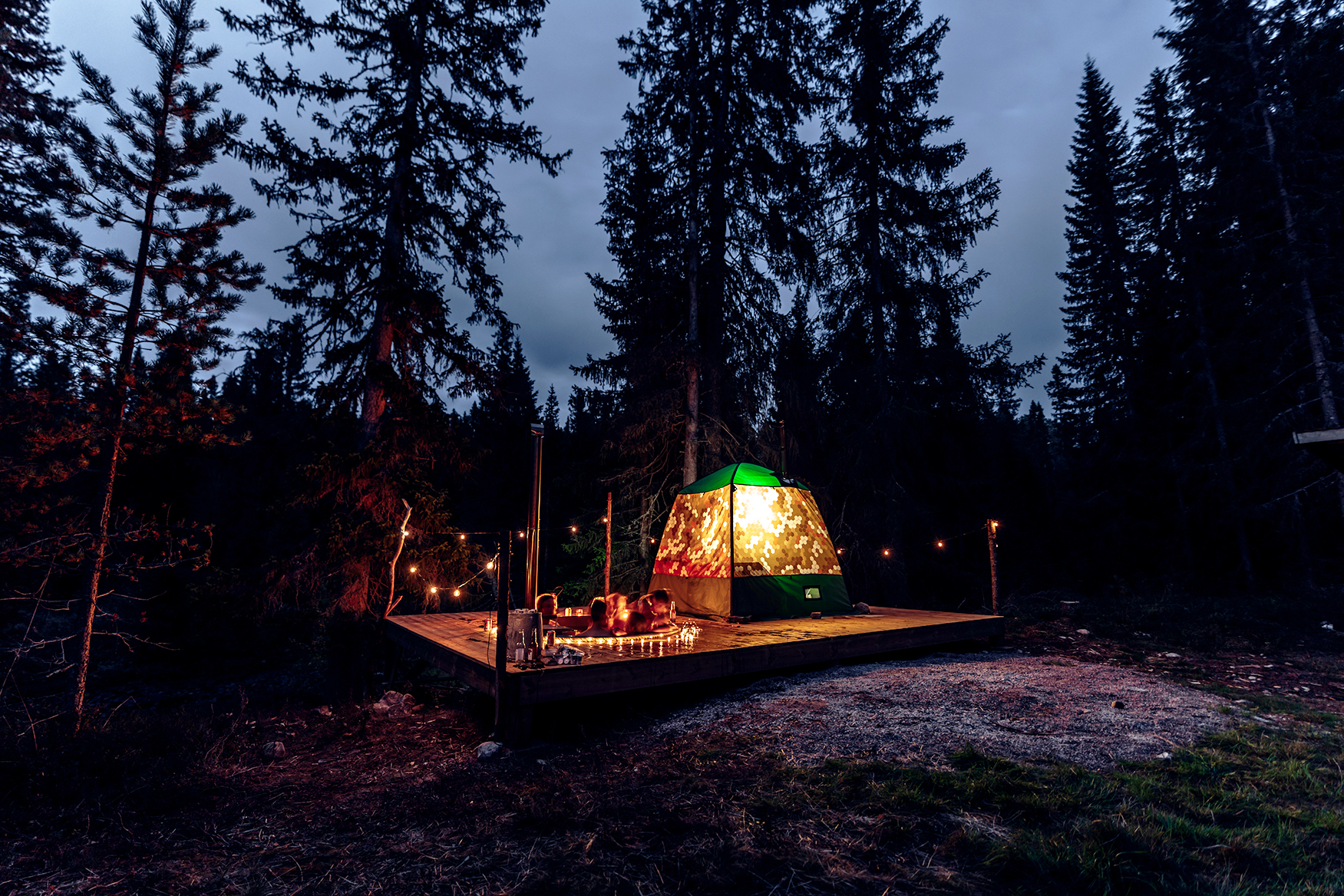 A lit tent on a wooden platform in a forest at night, with people gathered around a fire and string lights overhead.