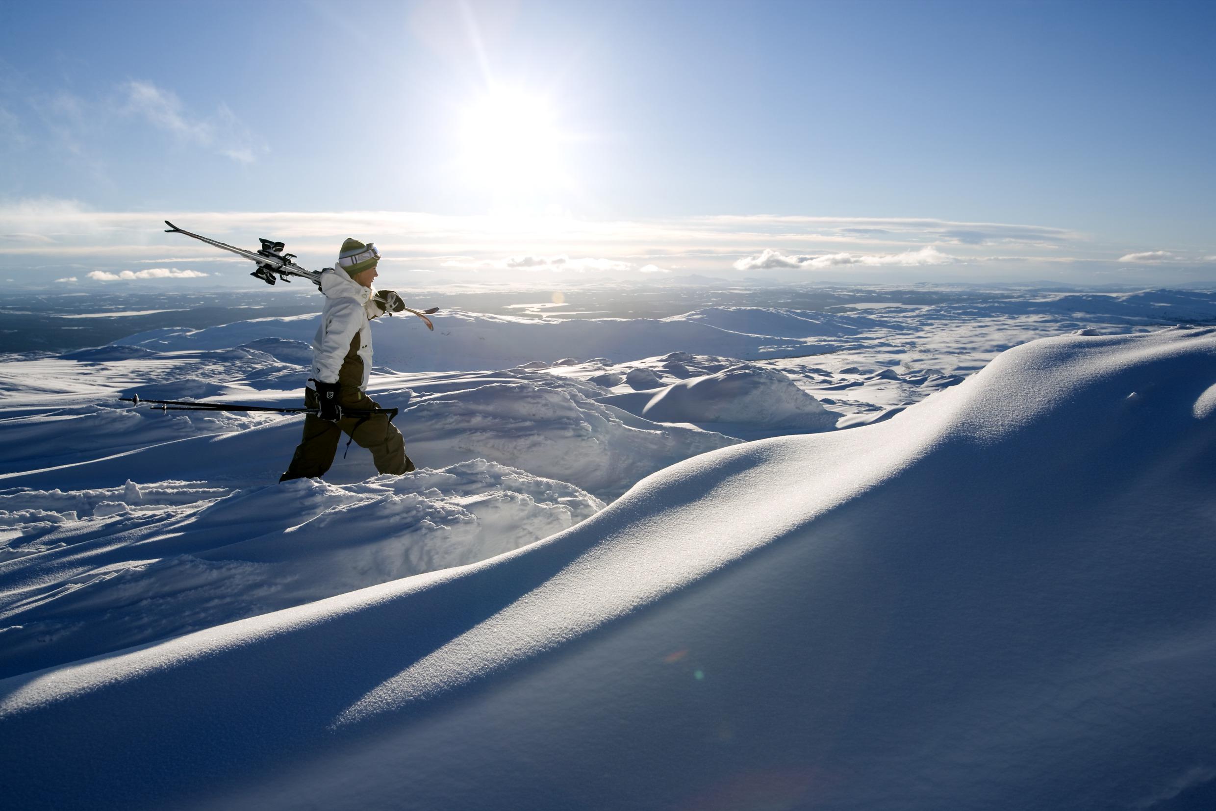 A man is walking through fresh powder snow on one of the Åre mountain peaks, holding his skis against his shoulder.