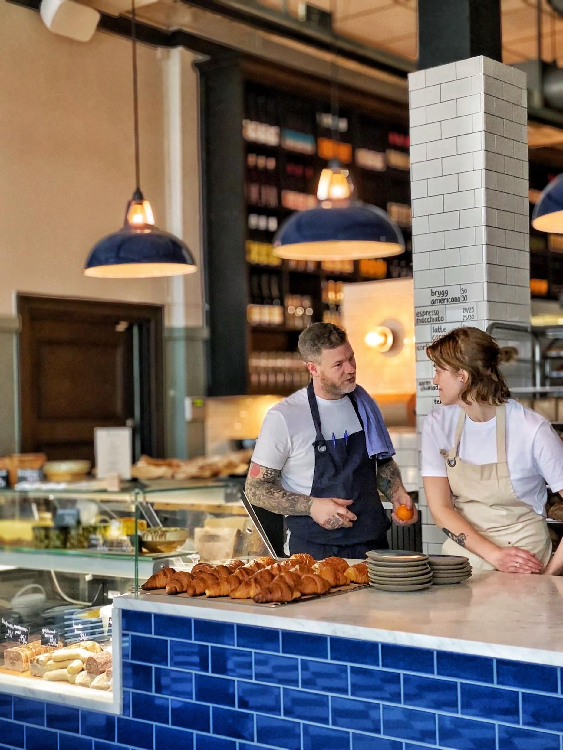 Twee obers praten in een café. Ze staan achter een toonbank met broodjes.