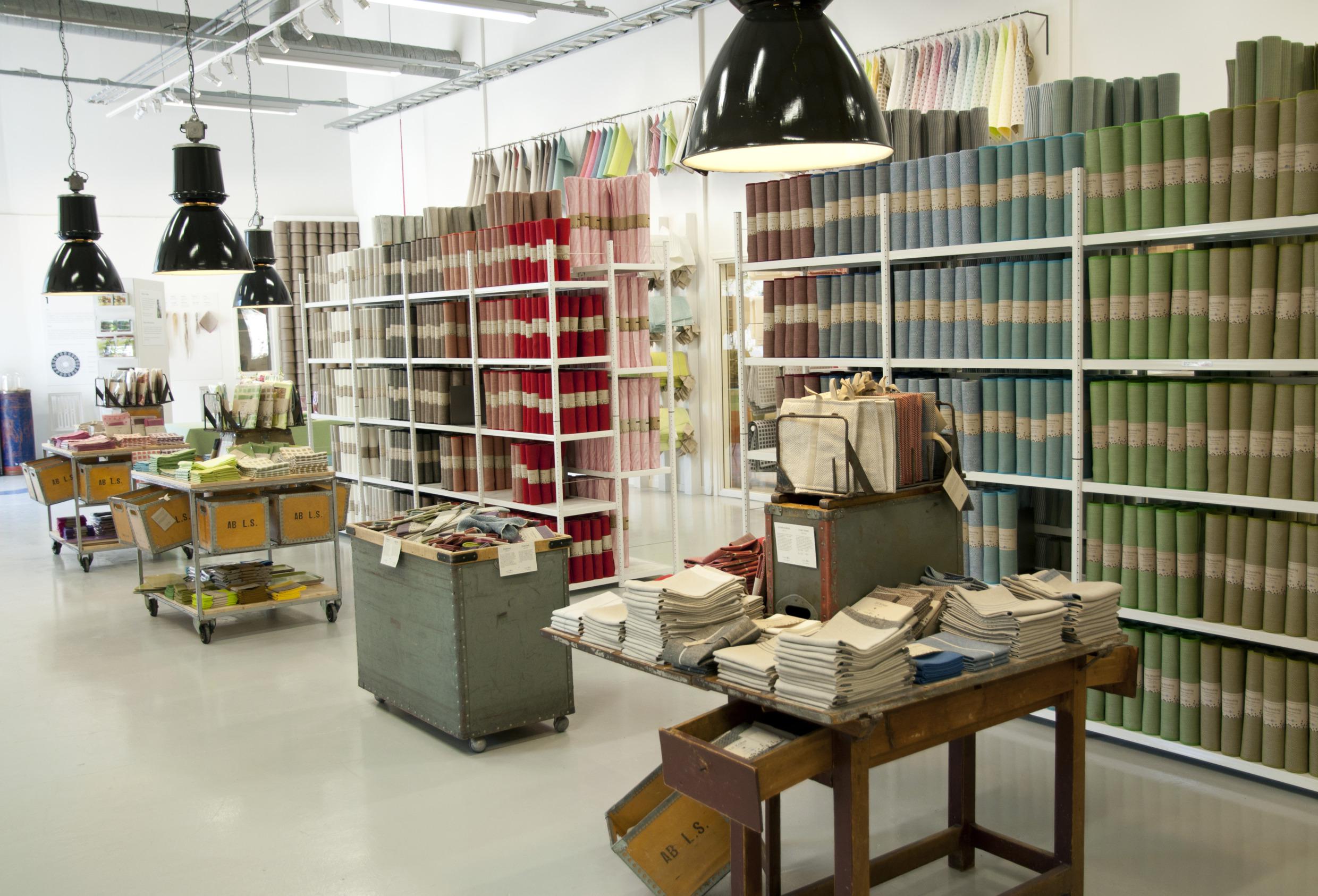 Des étagères avec des tissus en lin de différentes couleurs. Devant les étagères, il y a des tables avec des produits en lin.