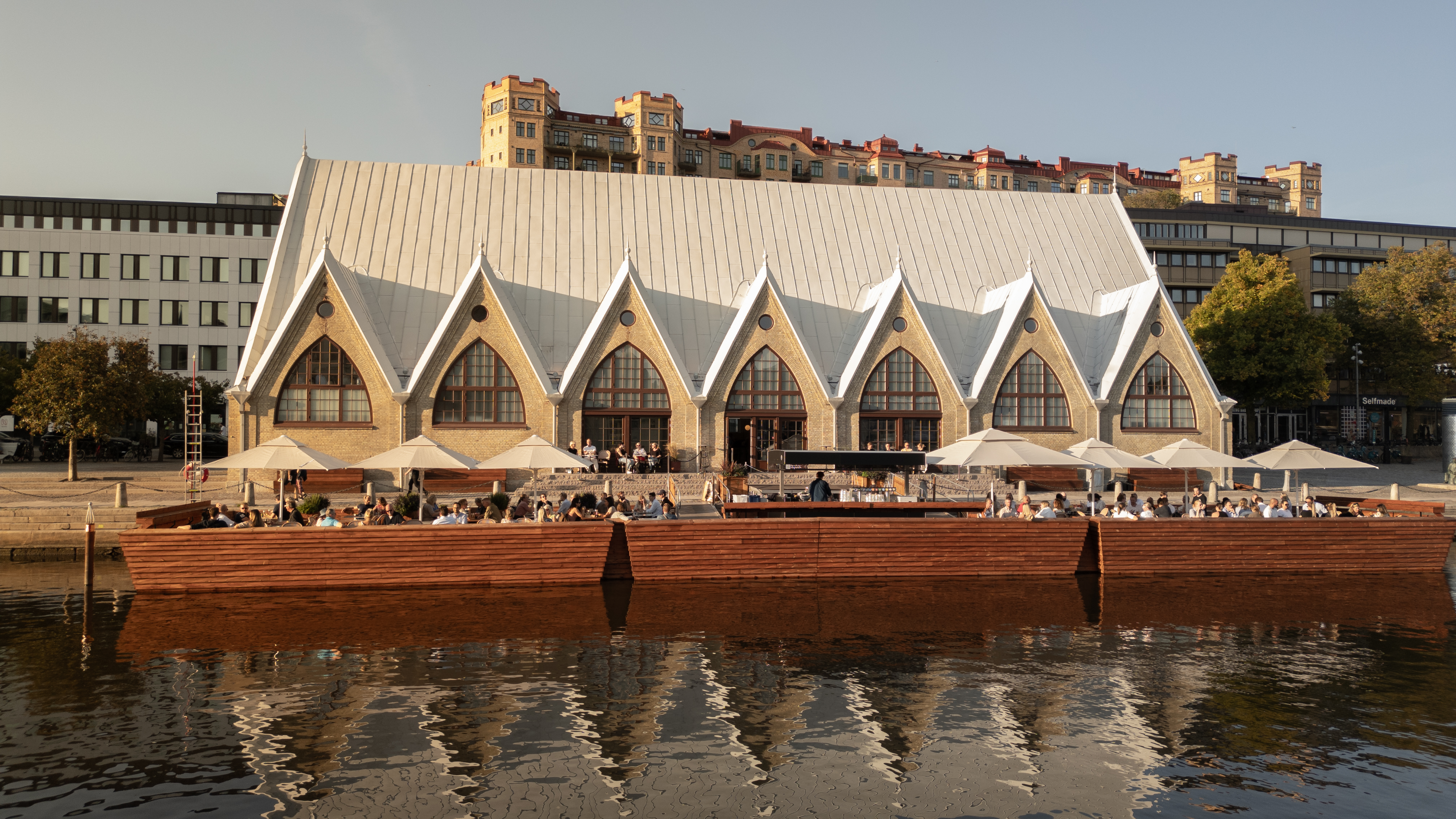 Der Fischmarkt Feskekörka in Göteborg, an einem sonnigen Tag vom anderen Ufer aus gesehen, mit Terrassen im Freien und Menschen, die am Wasser speisen.