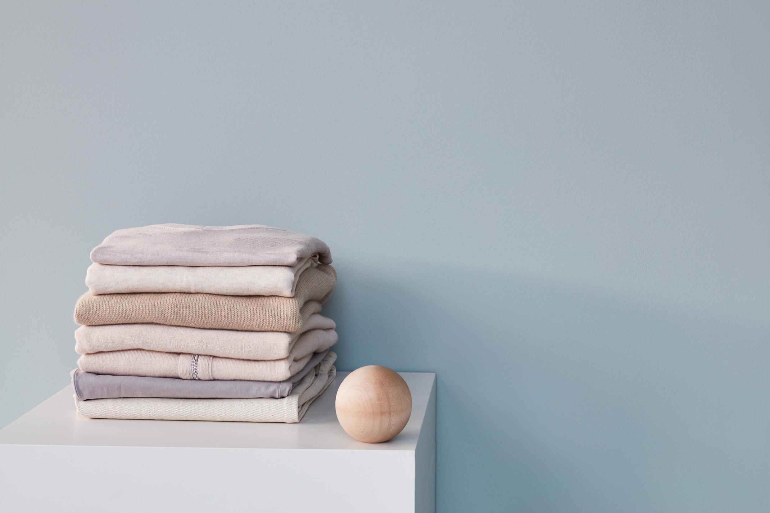 Des vêtements clairs sont pliés et superposés les uns sur les autres sur un table carrée se tenant le long d'un mur. A côté des vêtements, il y a une boule en bois.
