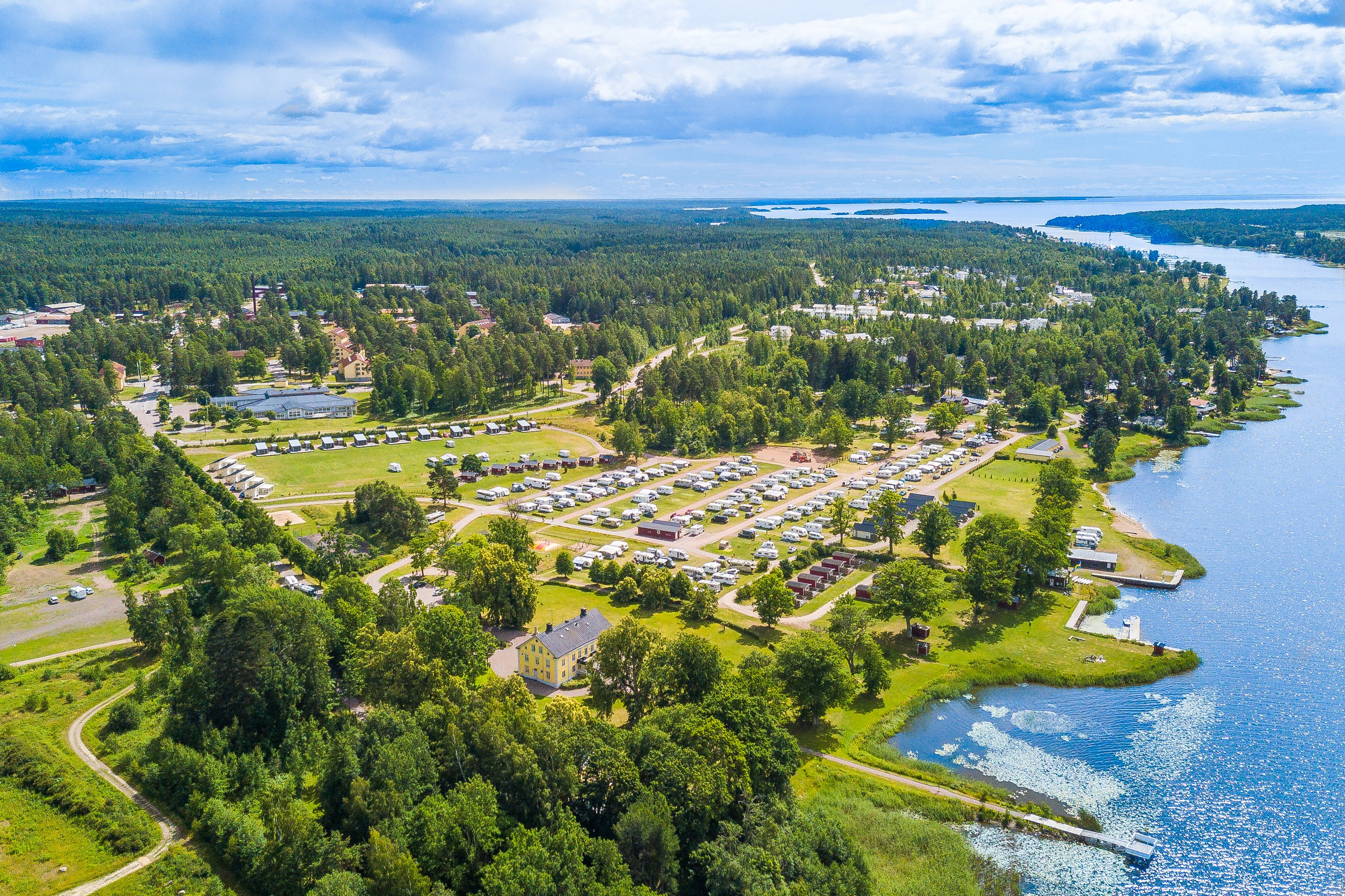 Aerial view of a lakeside campsite in Värmland with caravans, cabins and tents surrounded by forest and water.