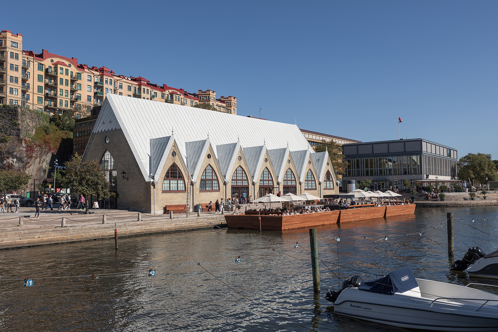 De vismarkt Feskekörka in Göteborg, gezien vanaf de overkant van het water op een zonnige dag, met terrassen en mensen die aan het water zitten te eten.