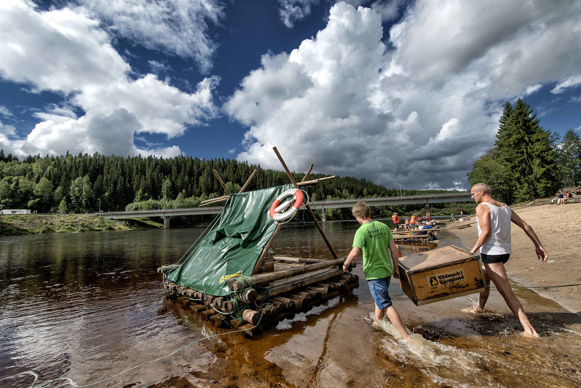 Two people carry equipment onto a timber raft in the Klarälven river in Värmland beneath dramatic clouds.