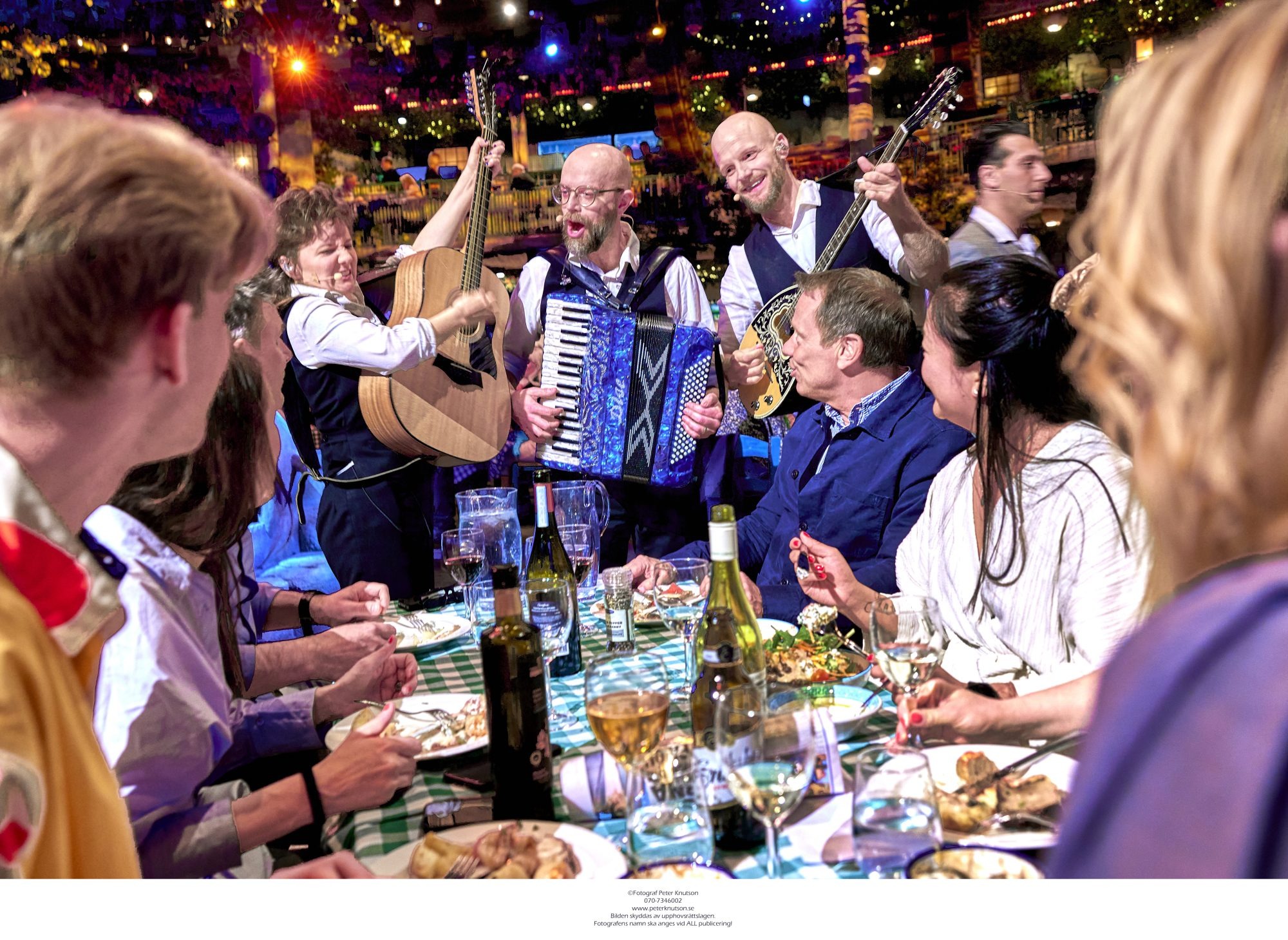 Drei Musiker singen und spielen Instrumente an einem Tisch, an dem Menschen zu Abend essen.