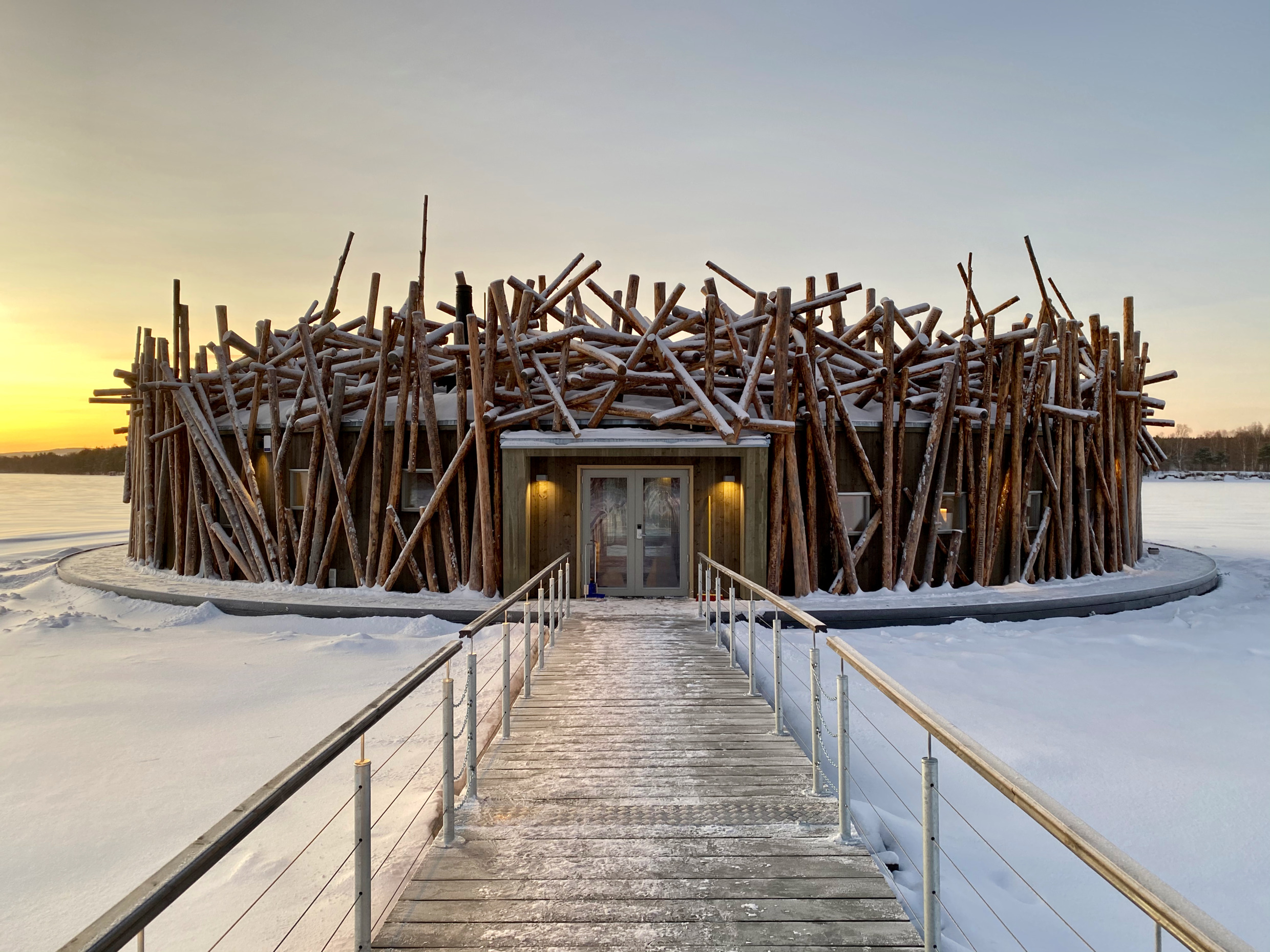 Eine Sauna, die im Winter auf einem schneebedeckten Fluss schwimmt. Die Außenseite der Sauna ist mit Holzstämmen verziert.