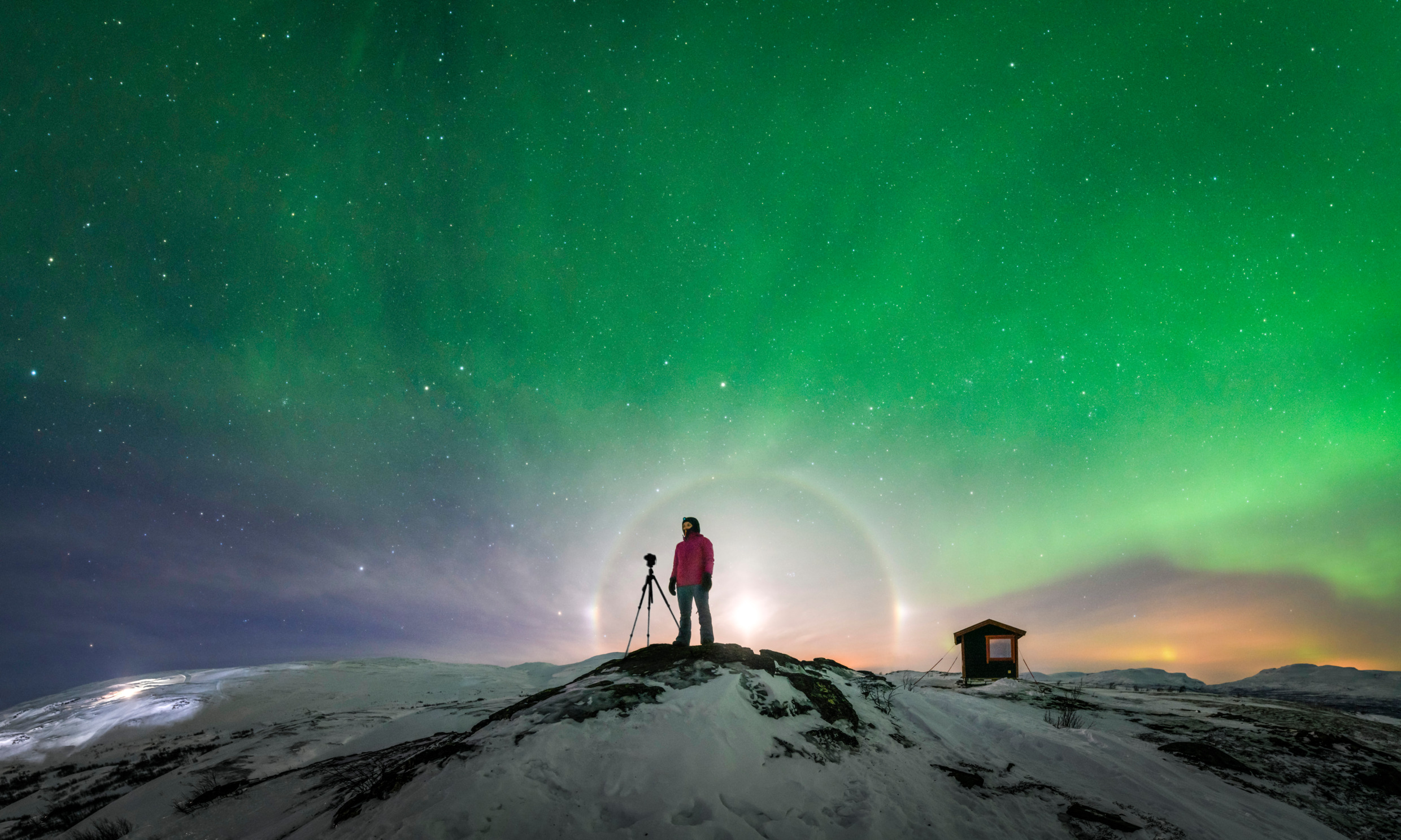 A person standing with its camera on a snowy hill under an epic aurora where the sky is changing colour from blue, yellow, purple, pink and a bright green.
