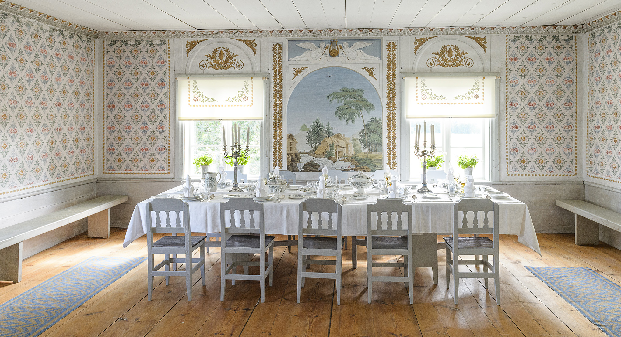 Dining room at Gästgivars Farmhouse with stencil-painted walls, long set table and mural of a rural scene.