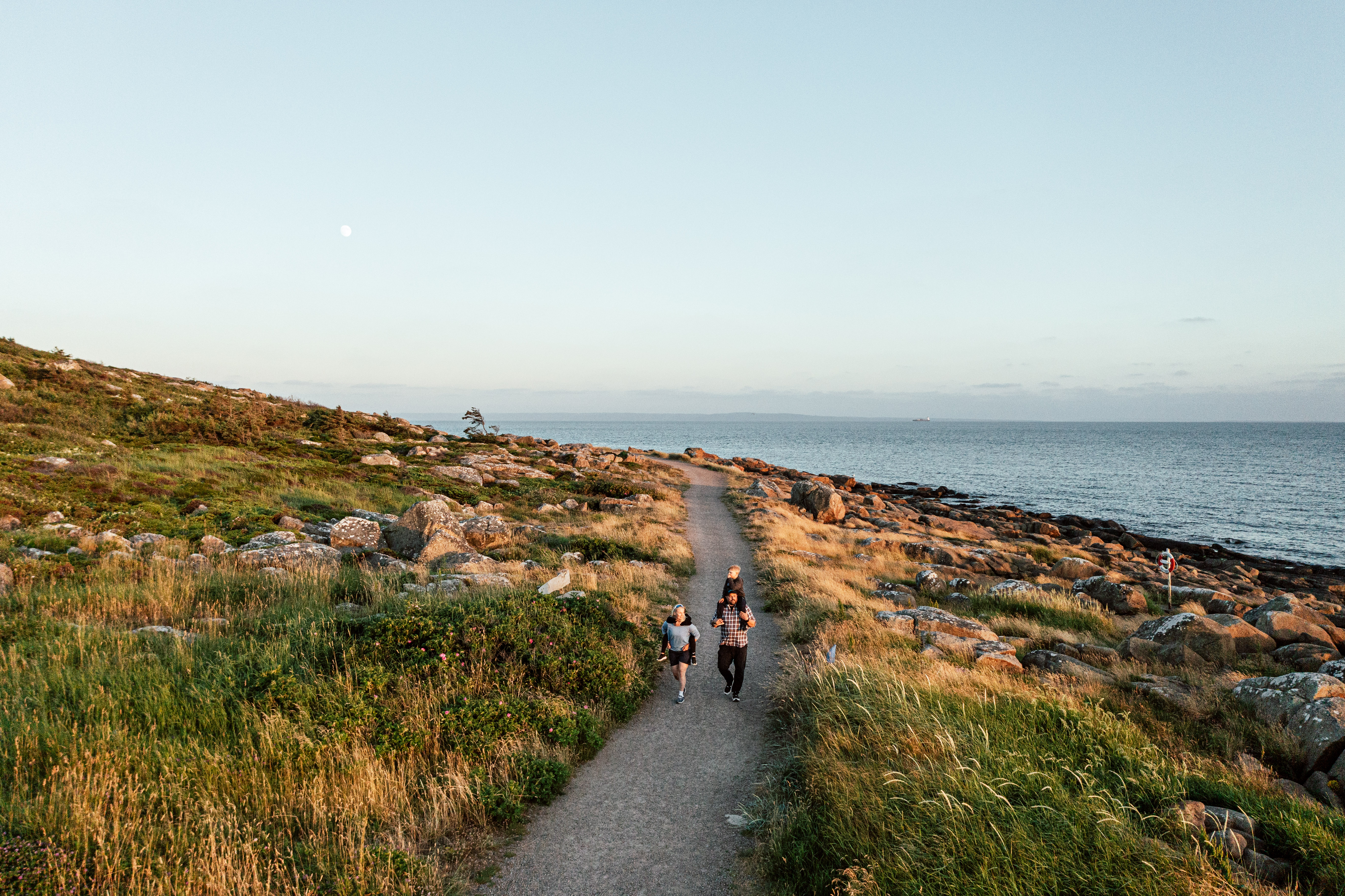 A family walking along a coastal path with grassy surroundings and rocky shoreline, with the sea in the background.
