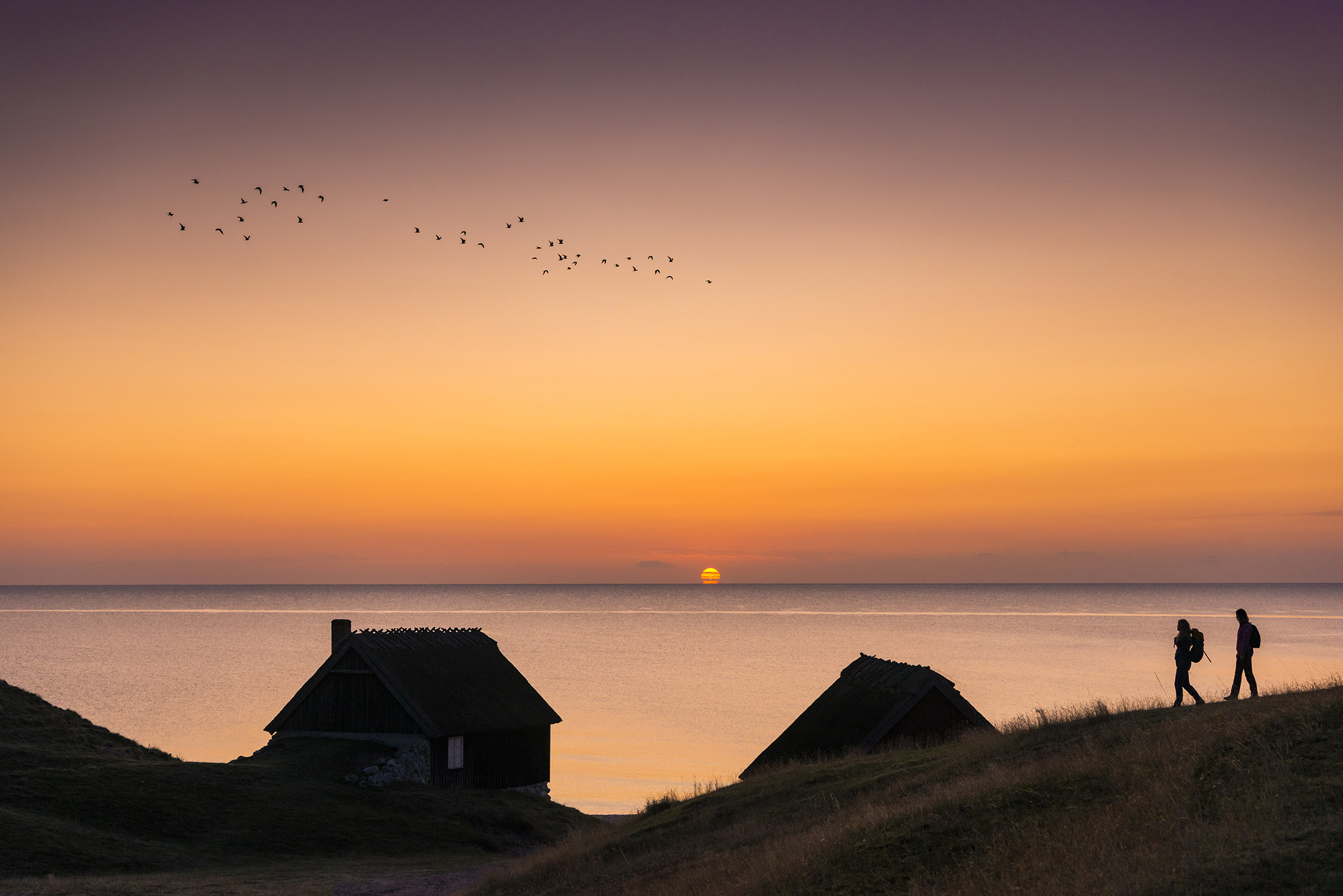 Zwei Wanderer, die bei Sonnenaufgang auf einem Küstenweg spazieren gehen, im Hintergrund kleine Hütten und das Meer.