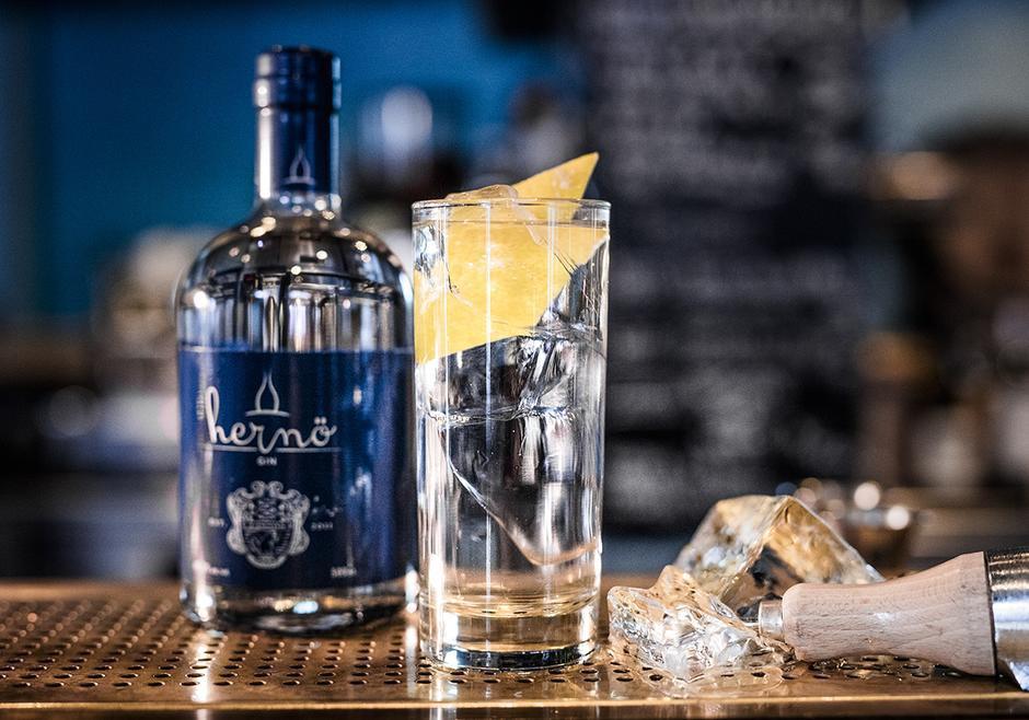 Eine Flasche Hernö Dry Gin steht neben einem Longdrinkglas mit Gin Tonic mit Eis.