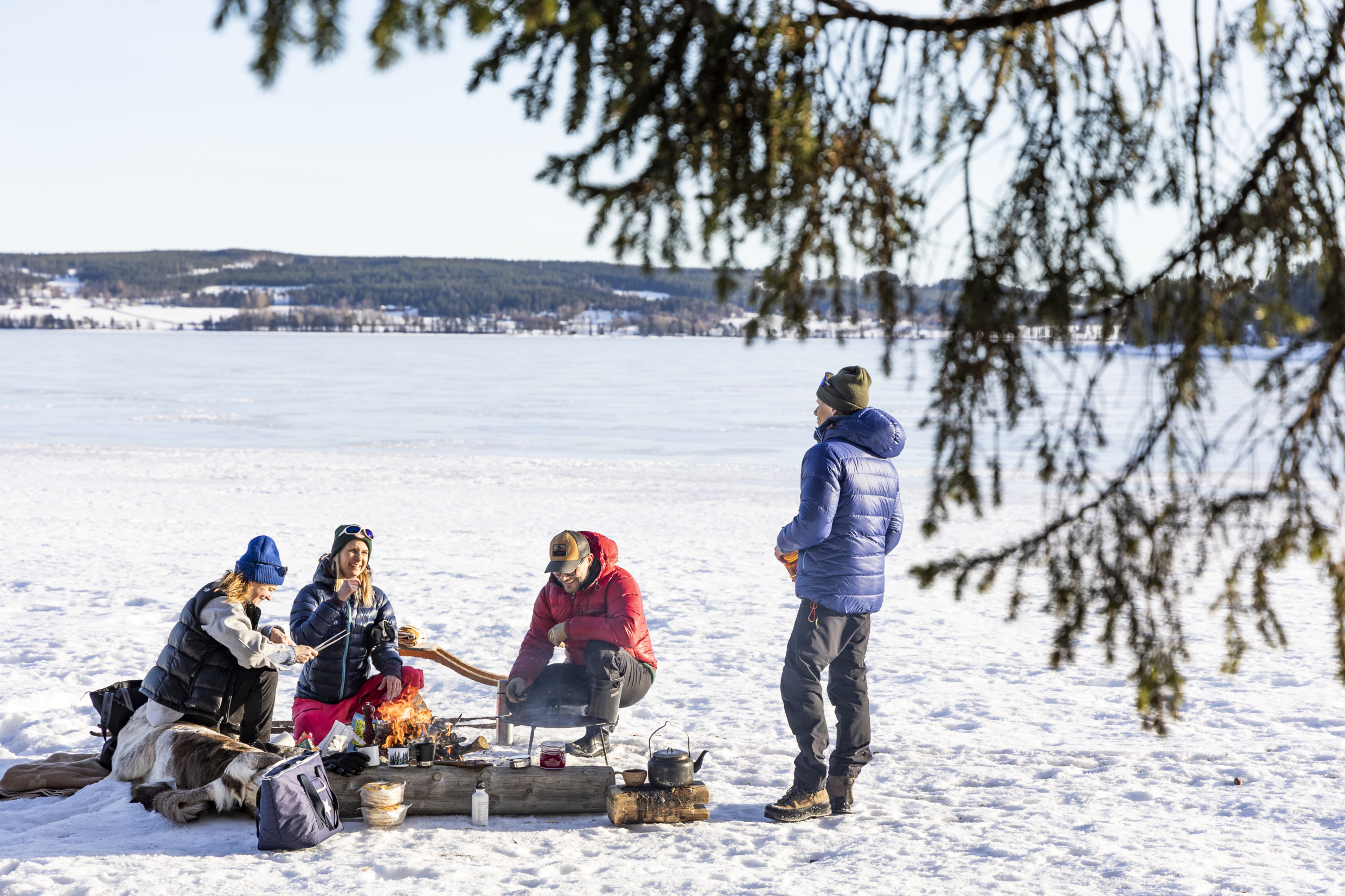 A group sits around a campfire on a sunny winter day having lunch.