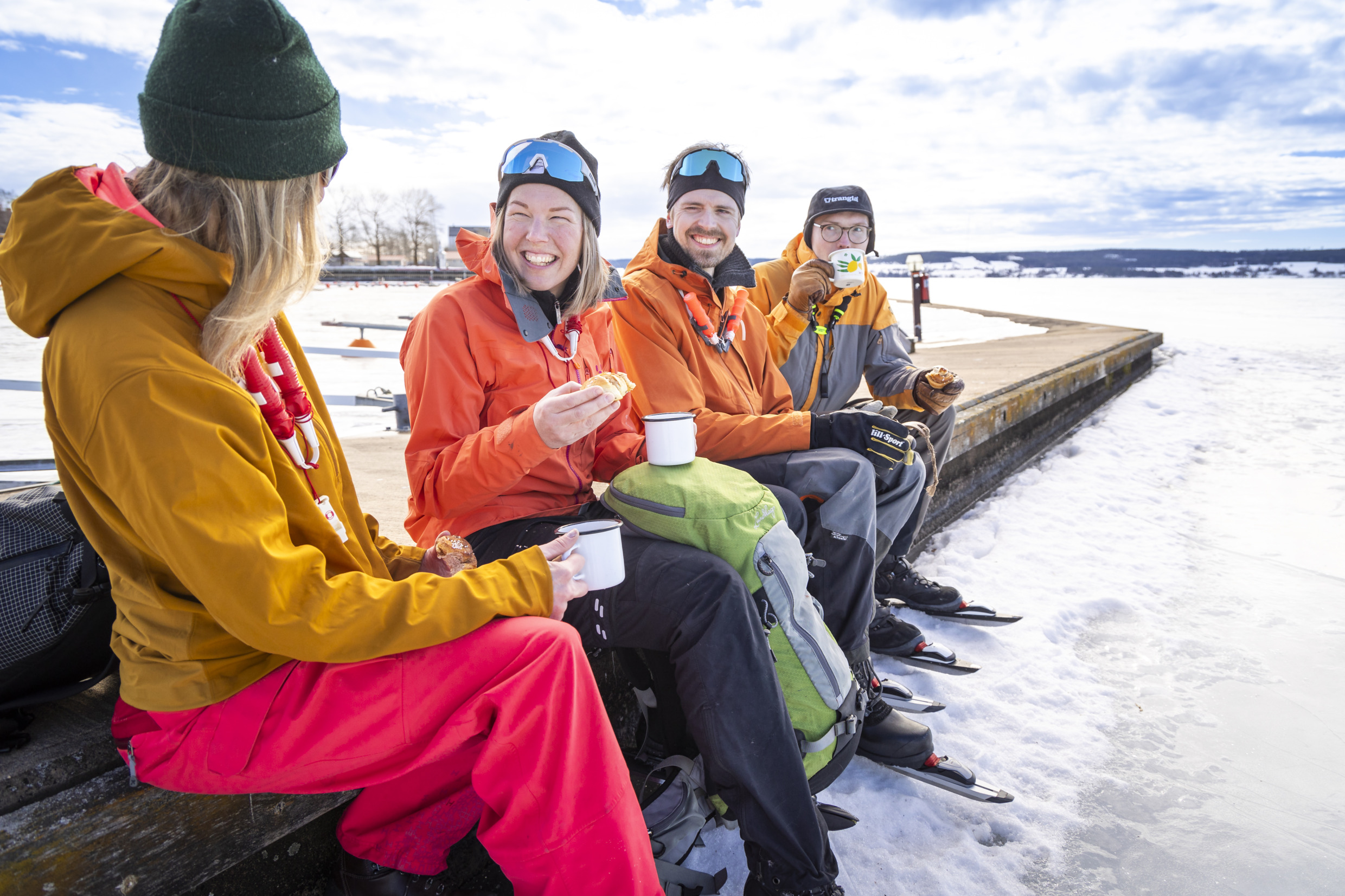 A group of four drinking coffee next to an ice-covered lake. They are wearing ice skates.
