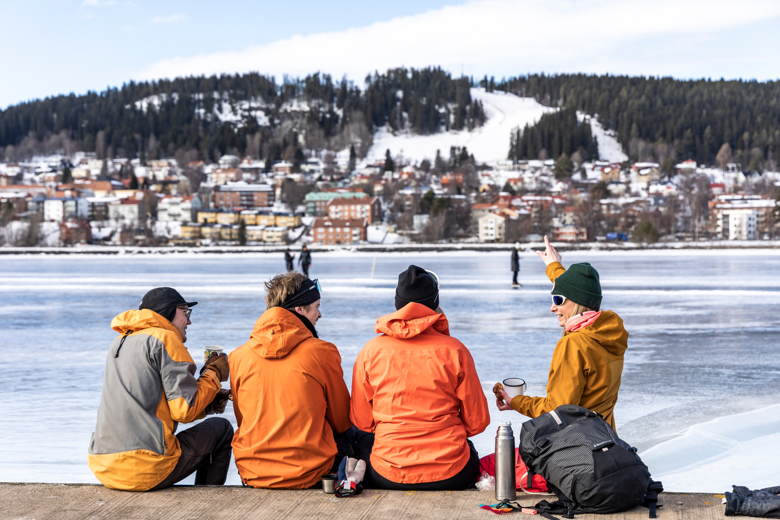 Eine Gruppe von vier Leuten sitzt neben einem zugefrorenen See, auf dem Leute Schlittschuh laufen. Die Gruppe trinkt Kaffee und einer von ihnen zeigt auf einen schneebedeckten Berg.