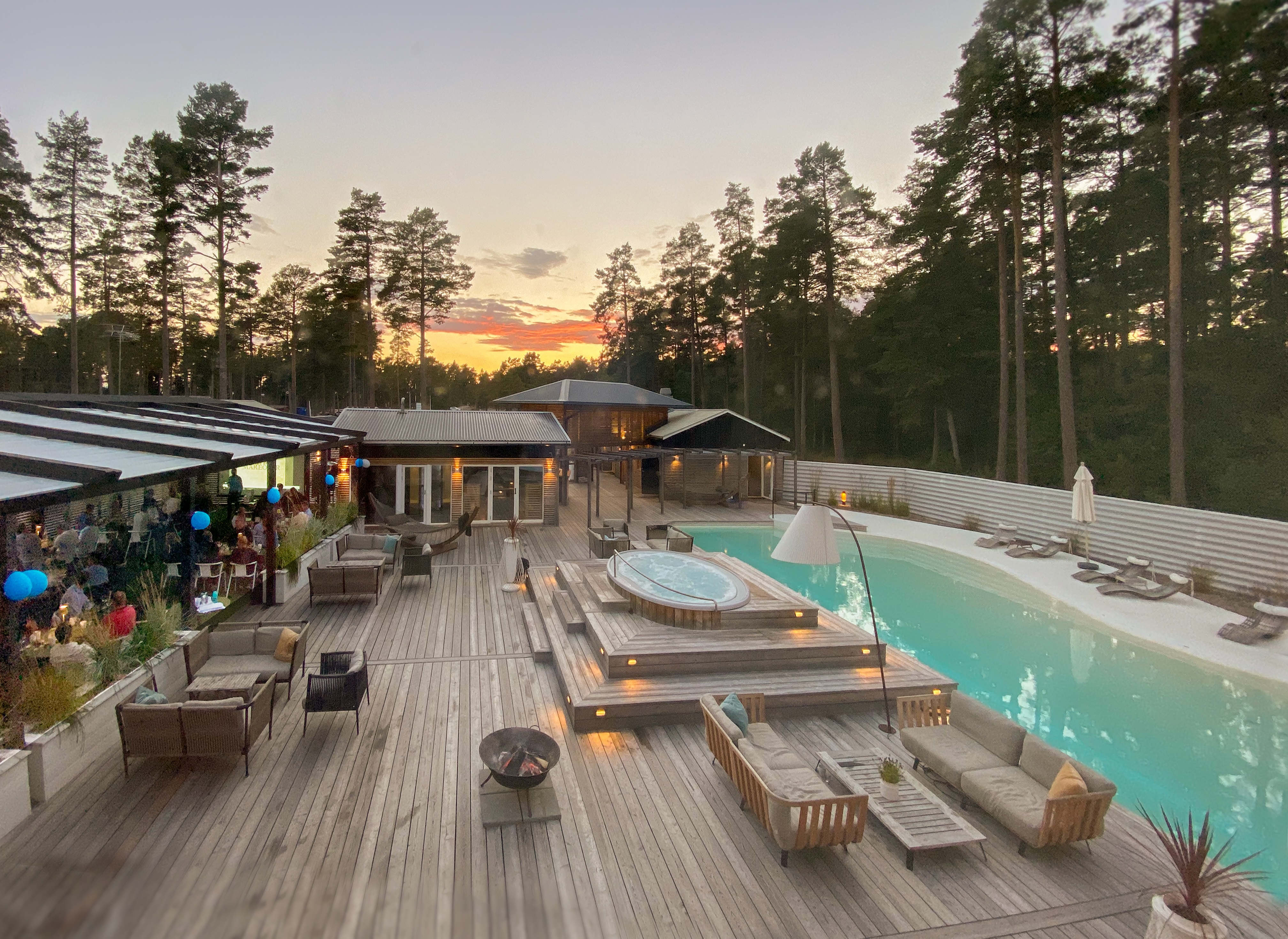 Outdoor pool area at Hölick Havsresort at sunset, with lounge furniture, jacuzzi, pine forest and guests dining on the terrace.