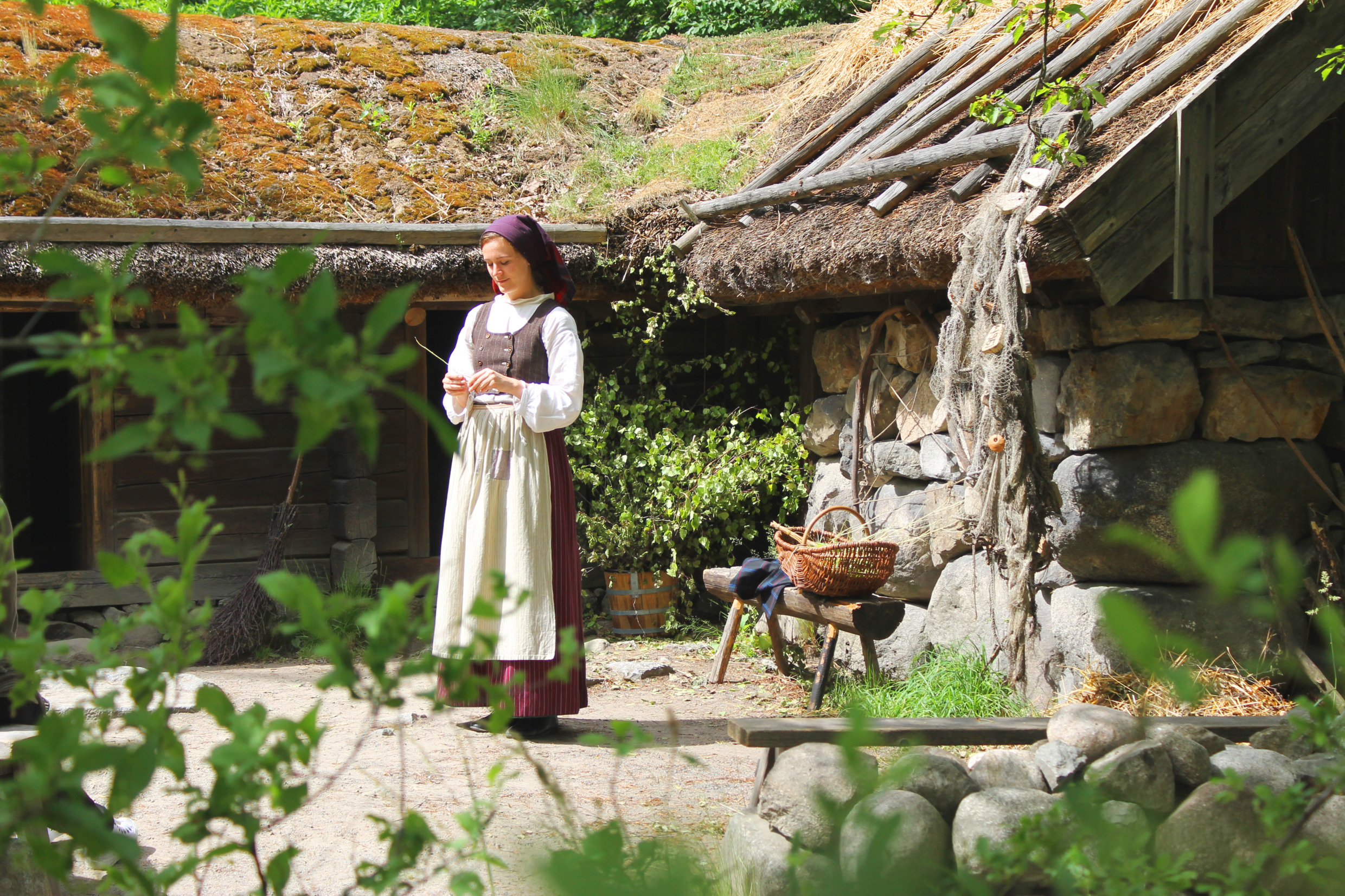 Woman in historical dress working with wool in front of a turf-roofed log cabin at Skansen, surrounded by greenery and rustic tools.