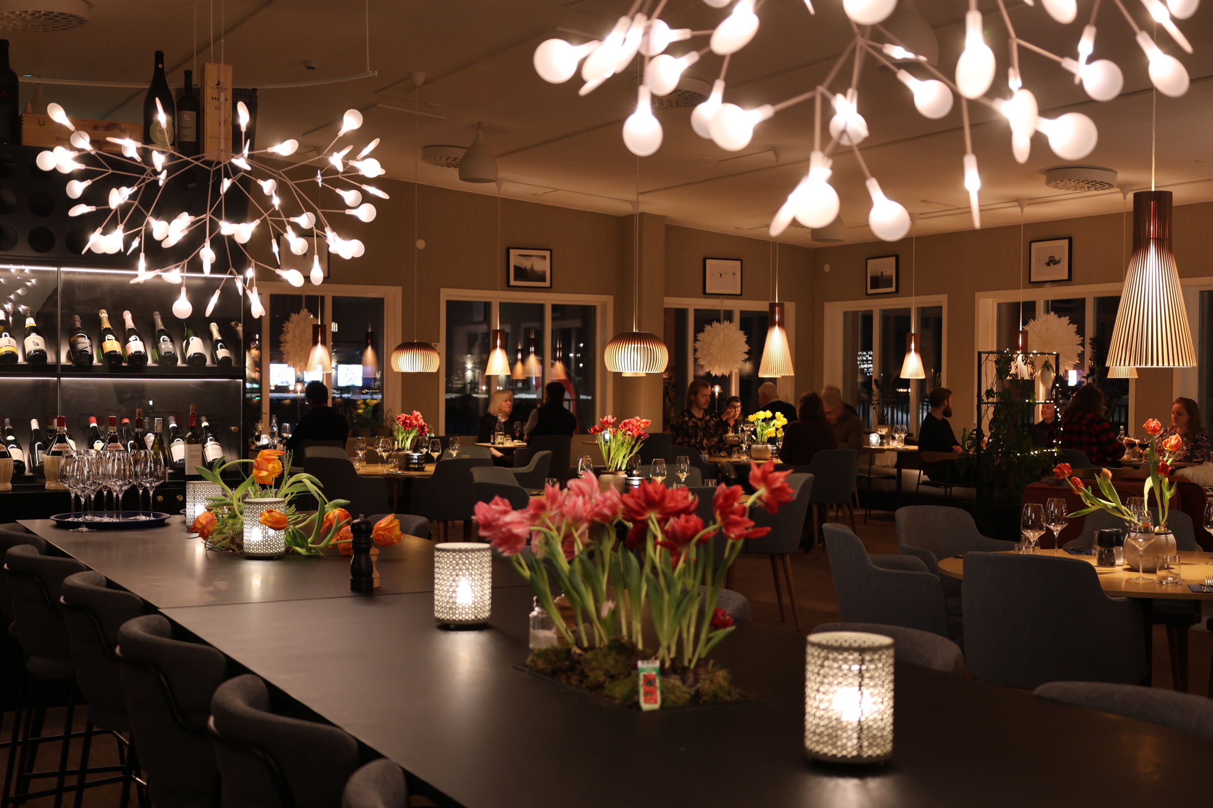 Een gezellig verlicht hotelrestaurant met bloemen op de tafels, wijnflessen en gasten die lekker eten in een relaxte sfeer.