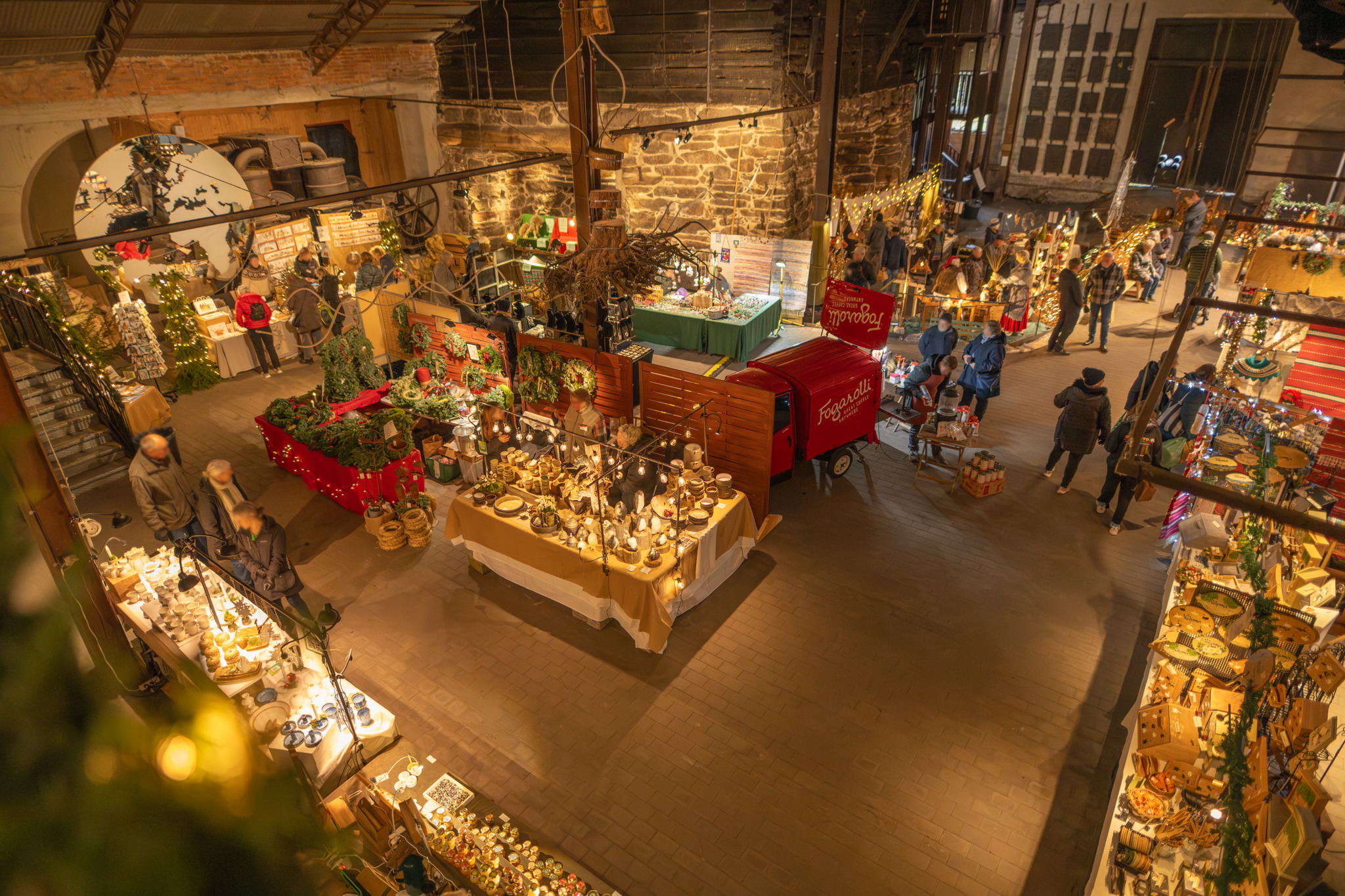 Vue intérieure du marché de Noël de Huseby Bruk avec ses étals, ses objets artisanaux, ses couronnes et ses visiteurs explorant le hall industriel historique.