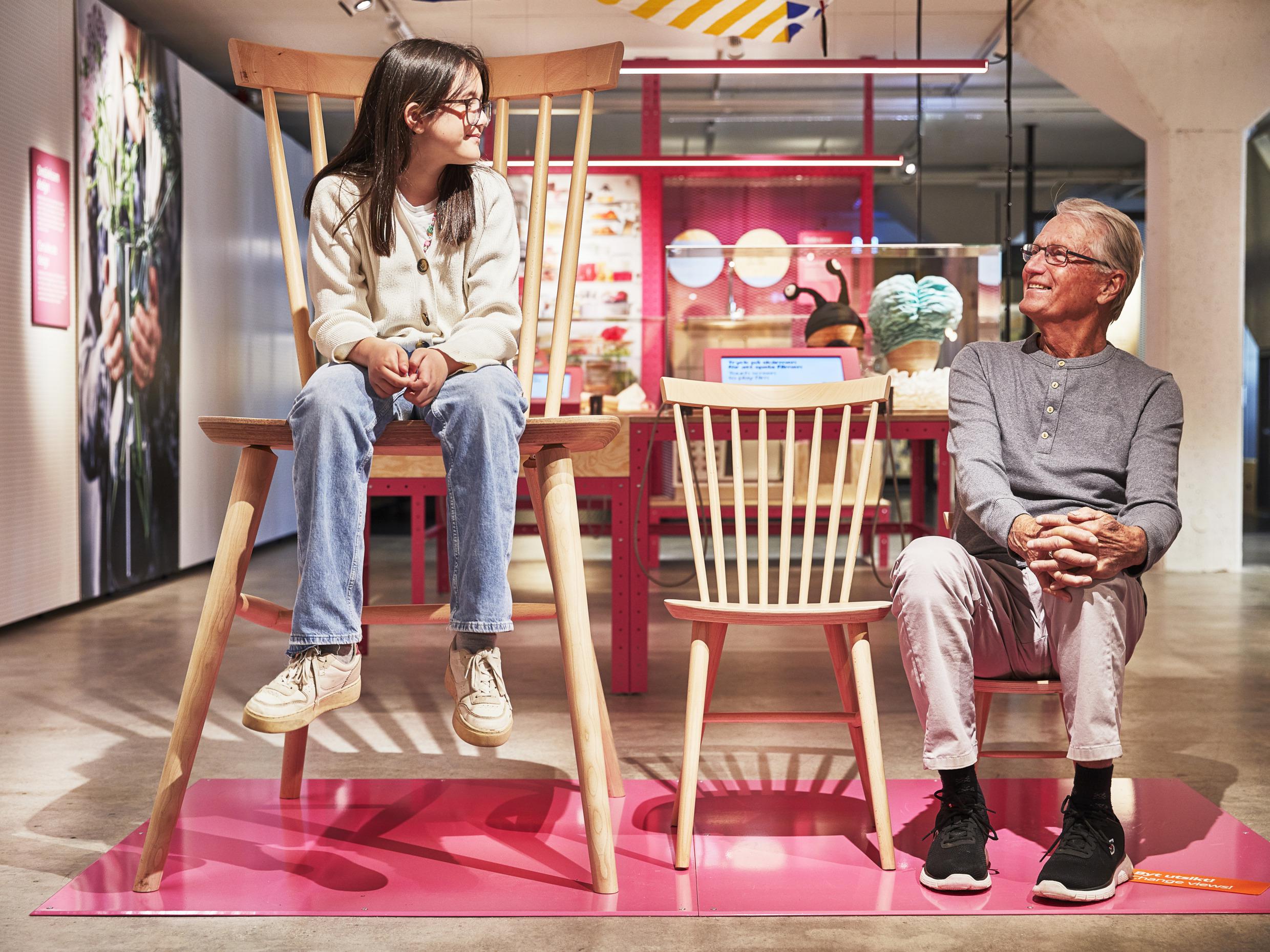 Drei Holzstühle in verschiedenen Größen. Auf dem größten Stuhl sitzt ein Kind und auf dem kleinsten Stuhl ein Mann.